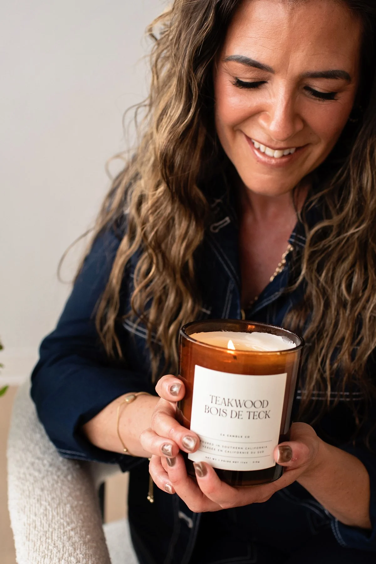 Chantal Menard smiling and holding a lit candle in a glass jar labeled 'Teakwood Bois de Teck'.