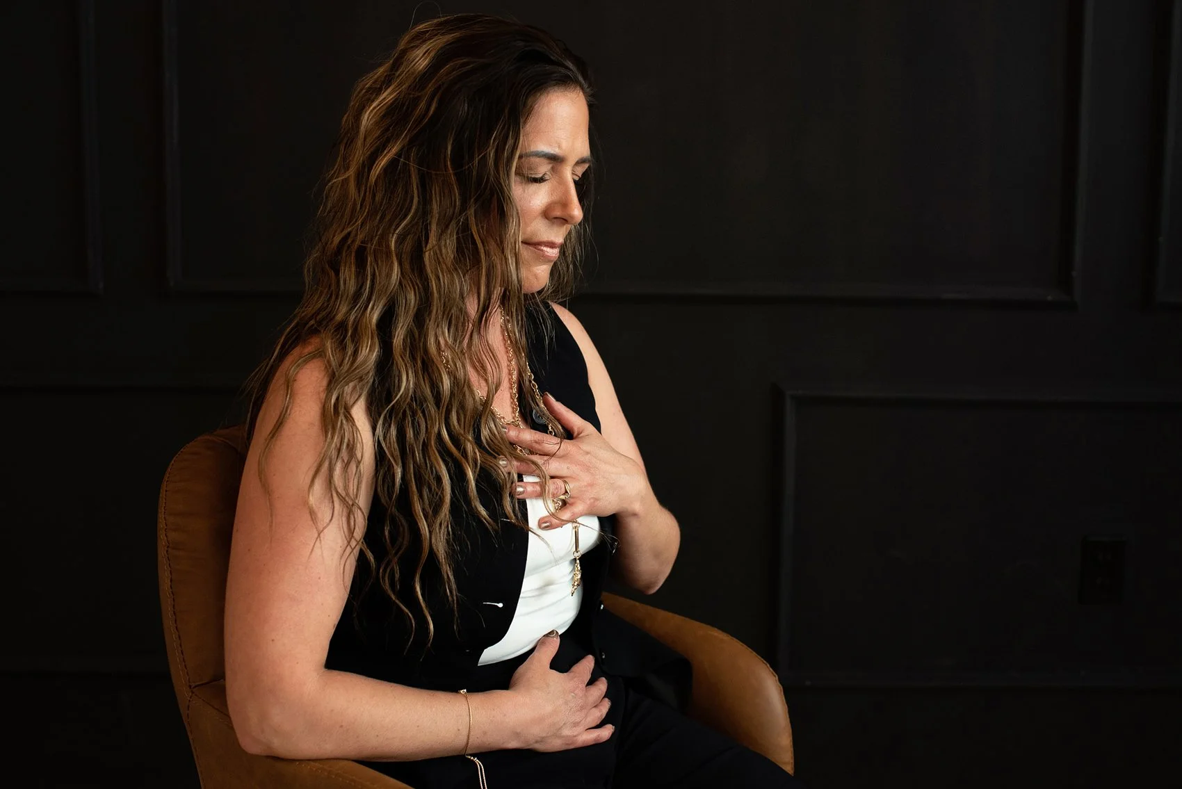 Chantal Menard sitting on a brown chair with her eyes closed and hand on her chest in a dark room with black walls.