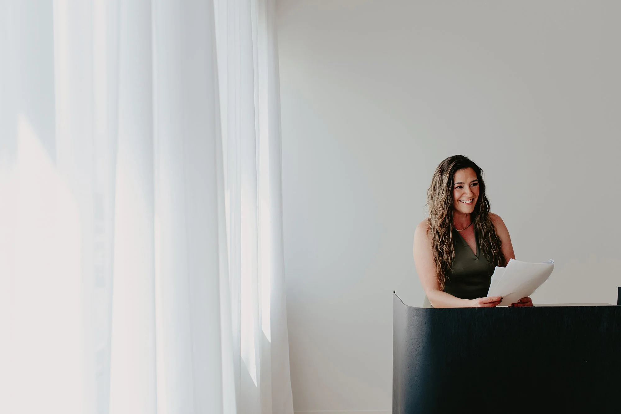 Chantal Menard smiling and reading papers while sitting at a desk in front of a window with white curtains.