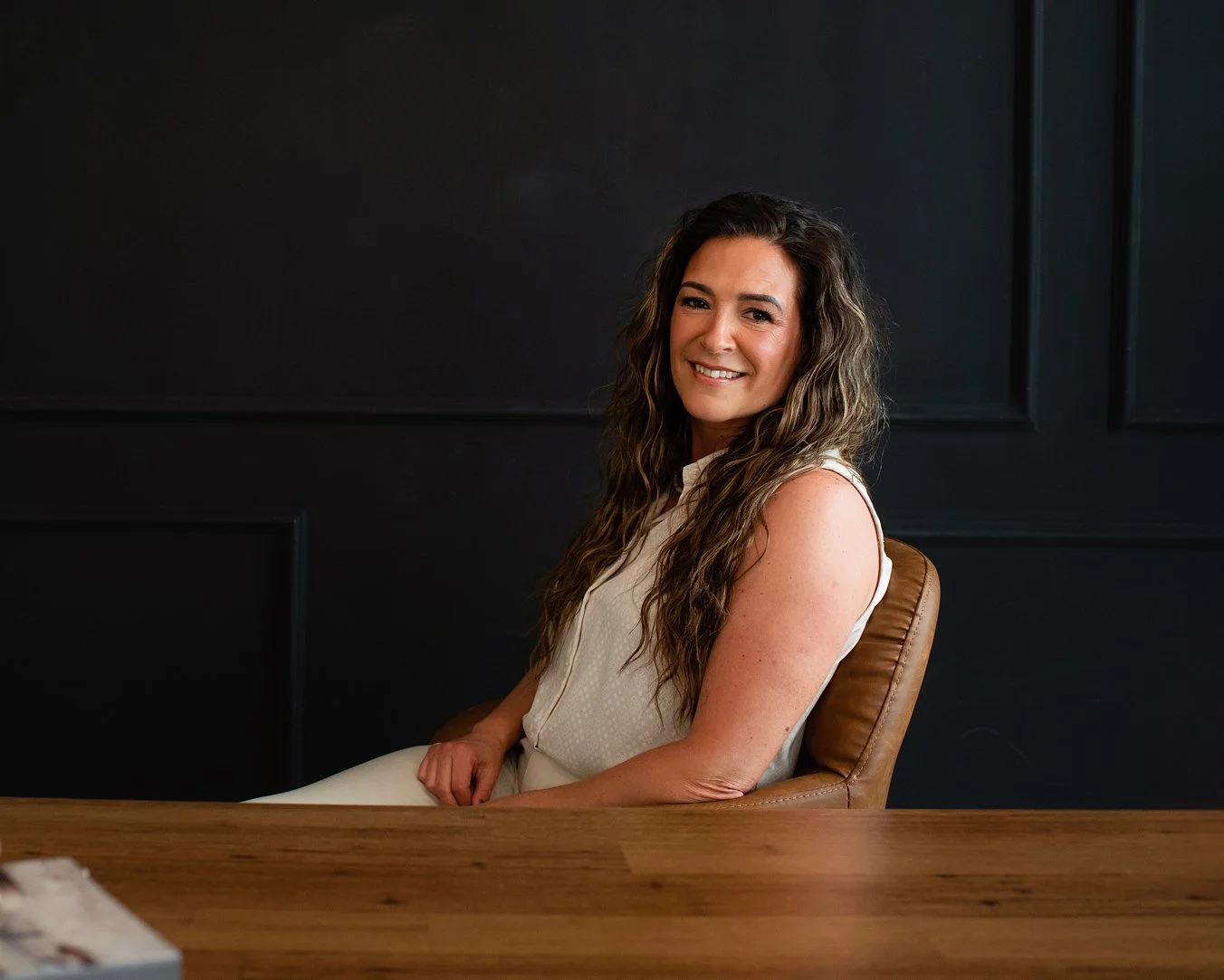 Chantal Menard sitting on a brown leather chair at a wooden table in front of a dark wall, smiling at the camera.