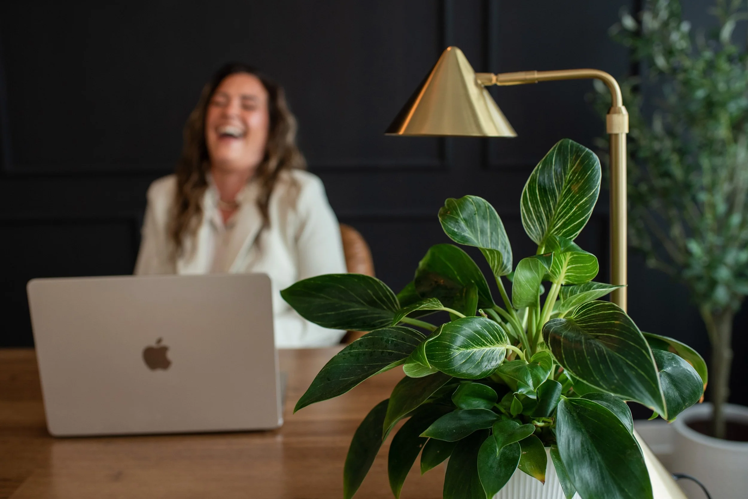 Chantal Menard laughing and working on a laptop at a desk with a large green potted plant in the foreground, a gold desk lamp, and a dark wall in the background.