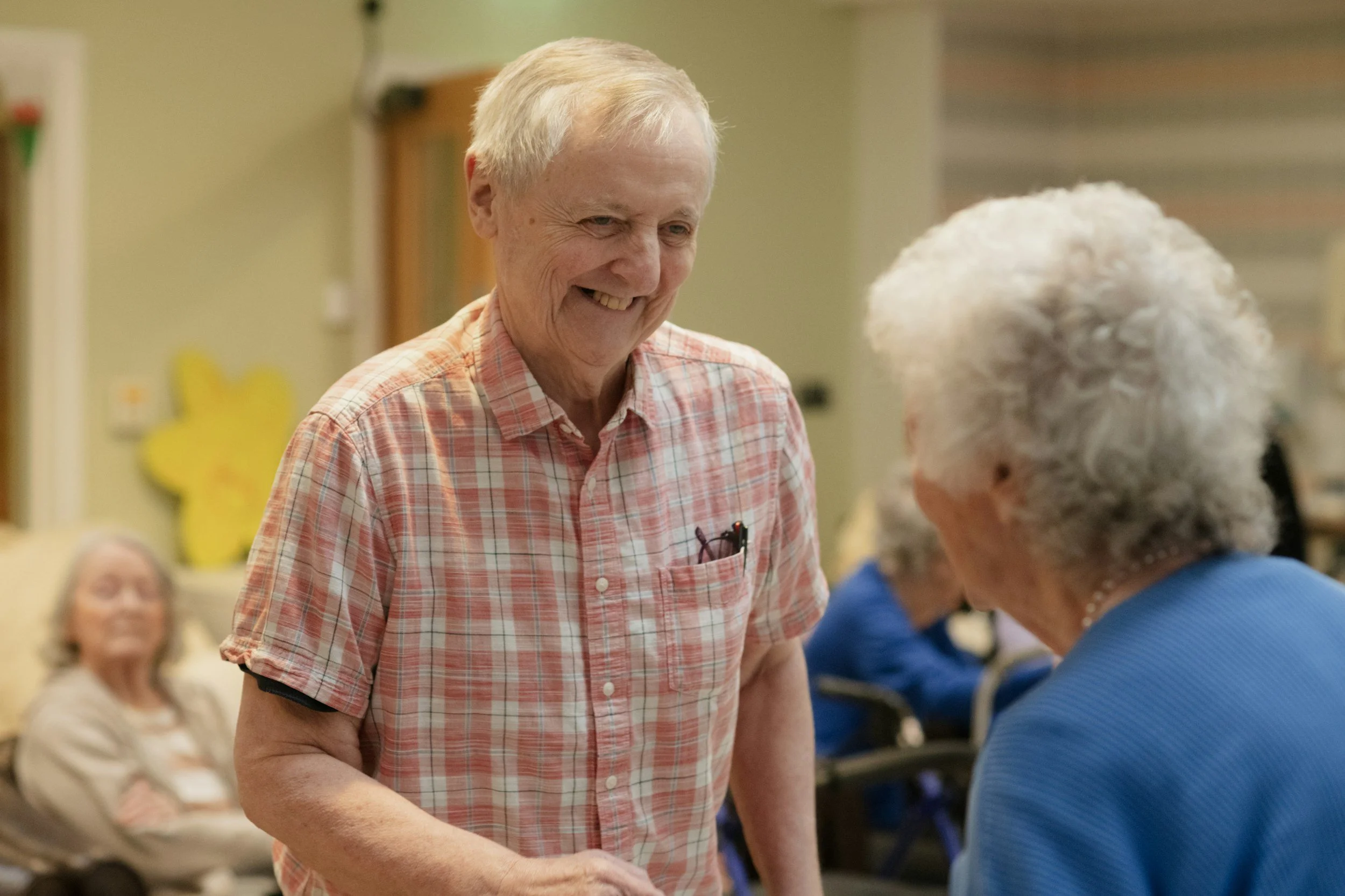 An elderly man with white hair, wearing a pink and white plaid short-sleeve shirt, is smiling and engaging in conversation with an elderly woman with curly white hair and a blue top, in a social gathering setting.