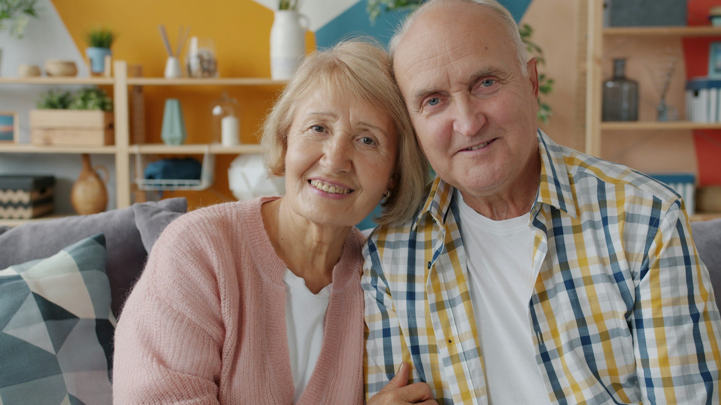 An elderly couple sitting close together on a sofa, smiling, with a colorful modern living room in the background.