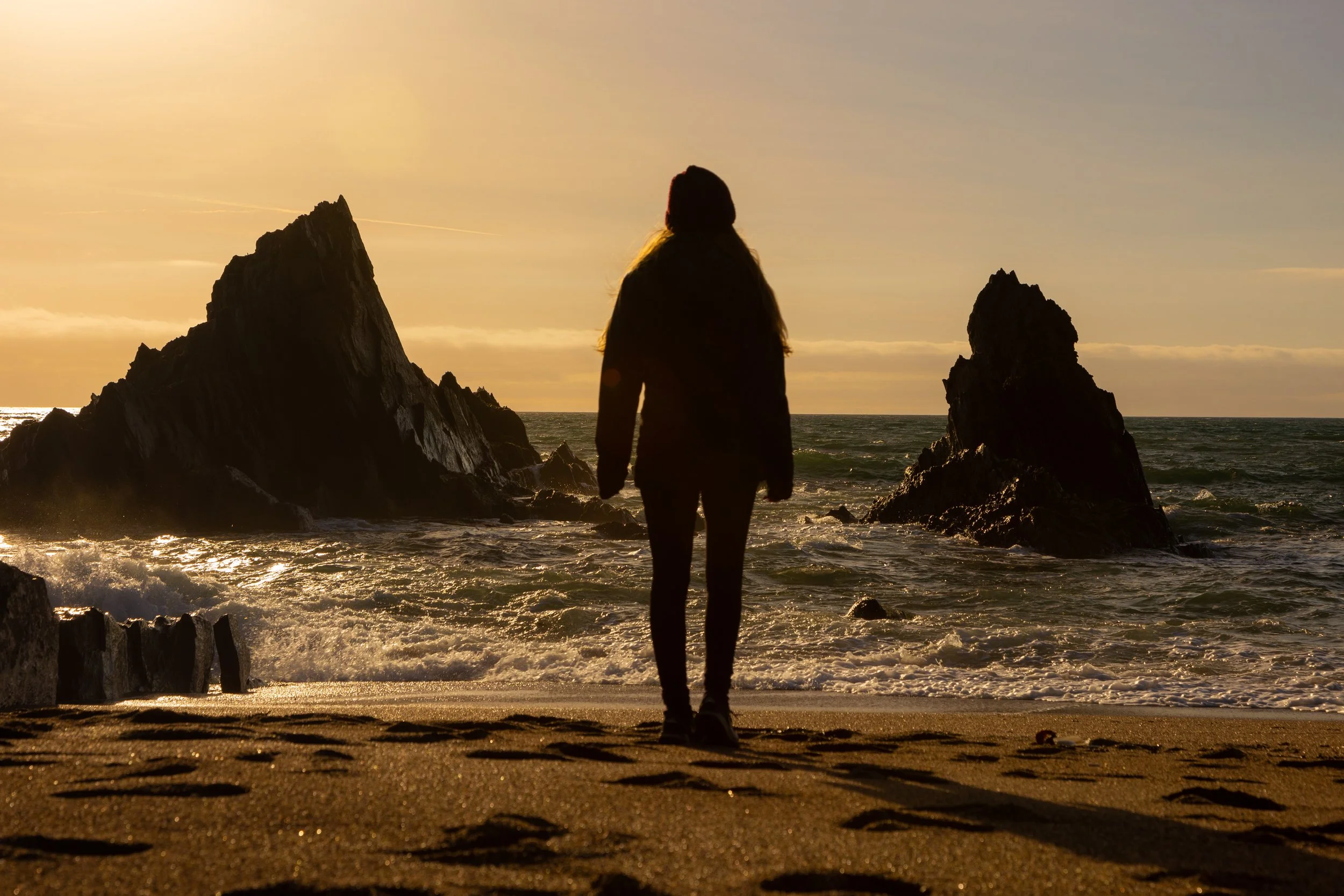 Person standing on the beach facing the ocean with rocky formations around during sunset.