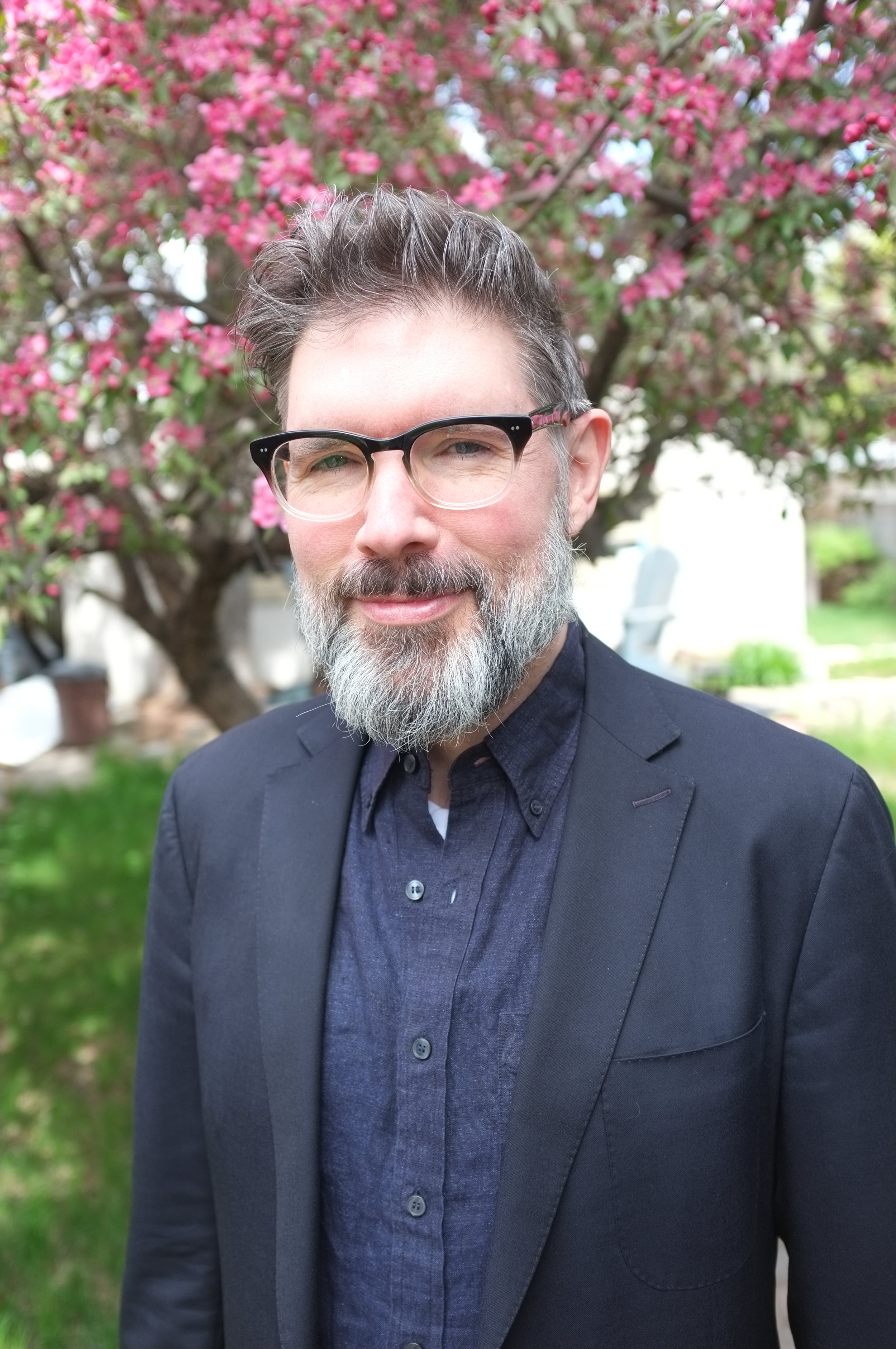 A man with glasses and a beard wearing a dark suit and shirt, standing outdoors in front of a flowering tree with pink blossoms.