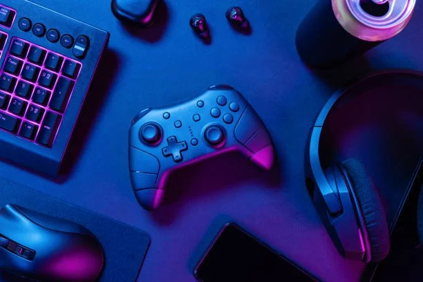 Gaming setup with a keyboard, game controller, wireless earbuds, a drink, a pair of headphones, and a mouse on a desk illuminated in blue and purple lighting.