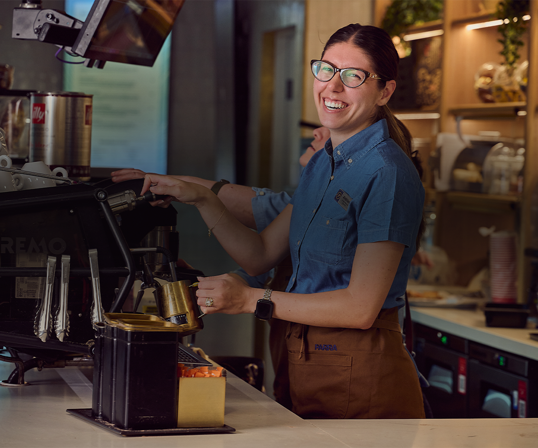 A woman barista wearing glasses and a blue shirt smiling as she prepares coffee behind a counter in a coffee shop.