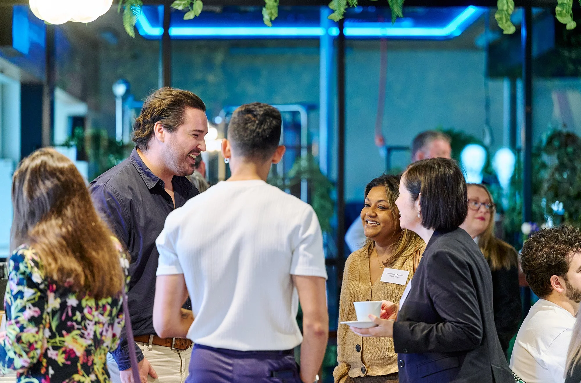 A group of diverse people socializing at a gathering, with some holding cups, in a modern, well-lit indoor space with greenery and blue accent lighting.