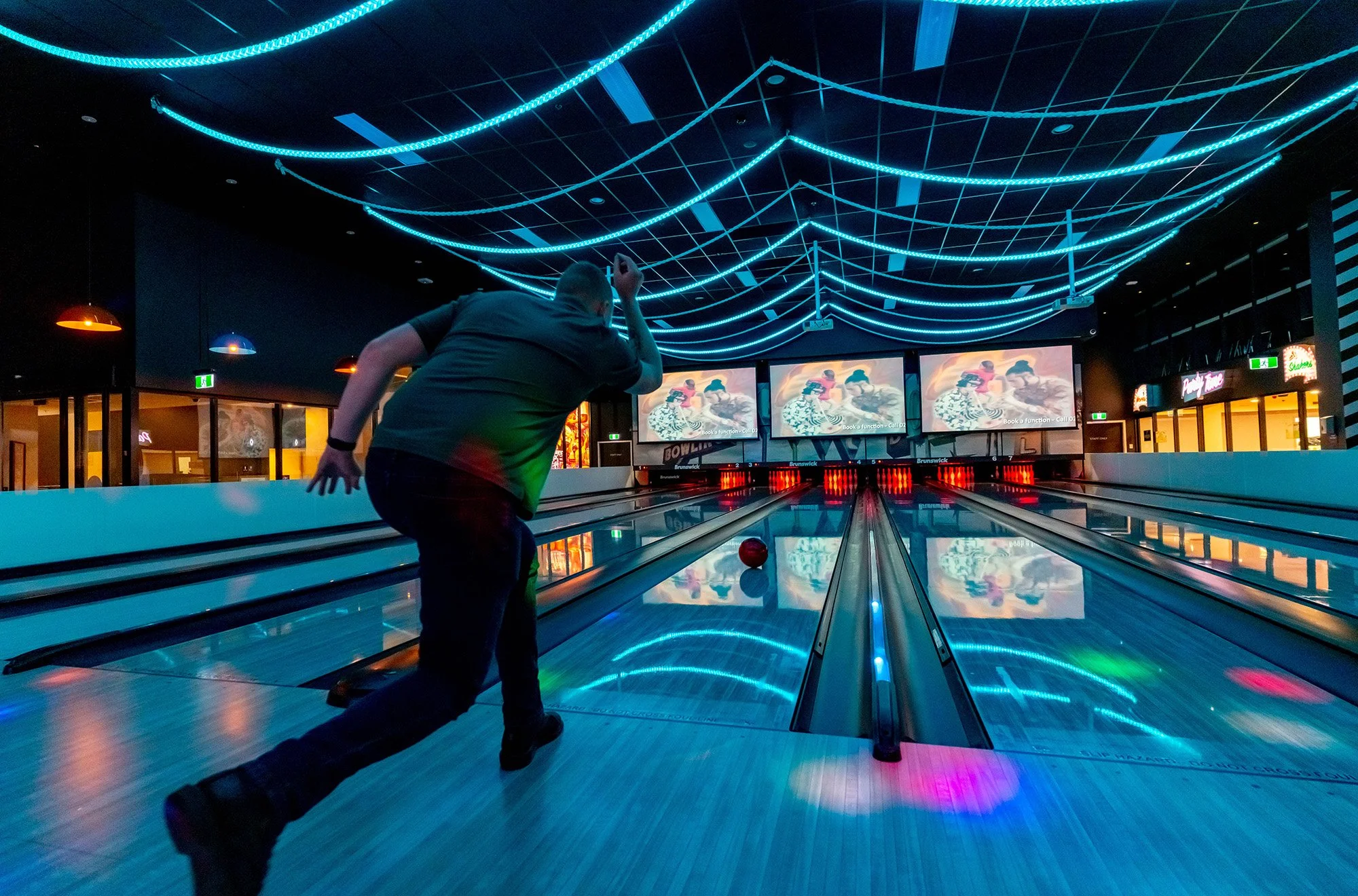 A person bowling in a brightly lit bowling alley with blue neon lights on the ceiling and multiple screens displaying images, as a bowling ball approaches the pins.