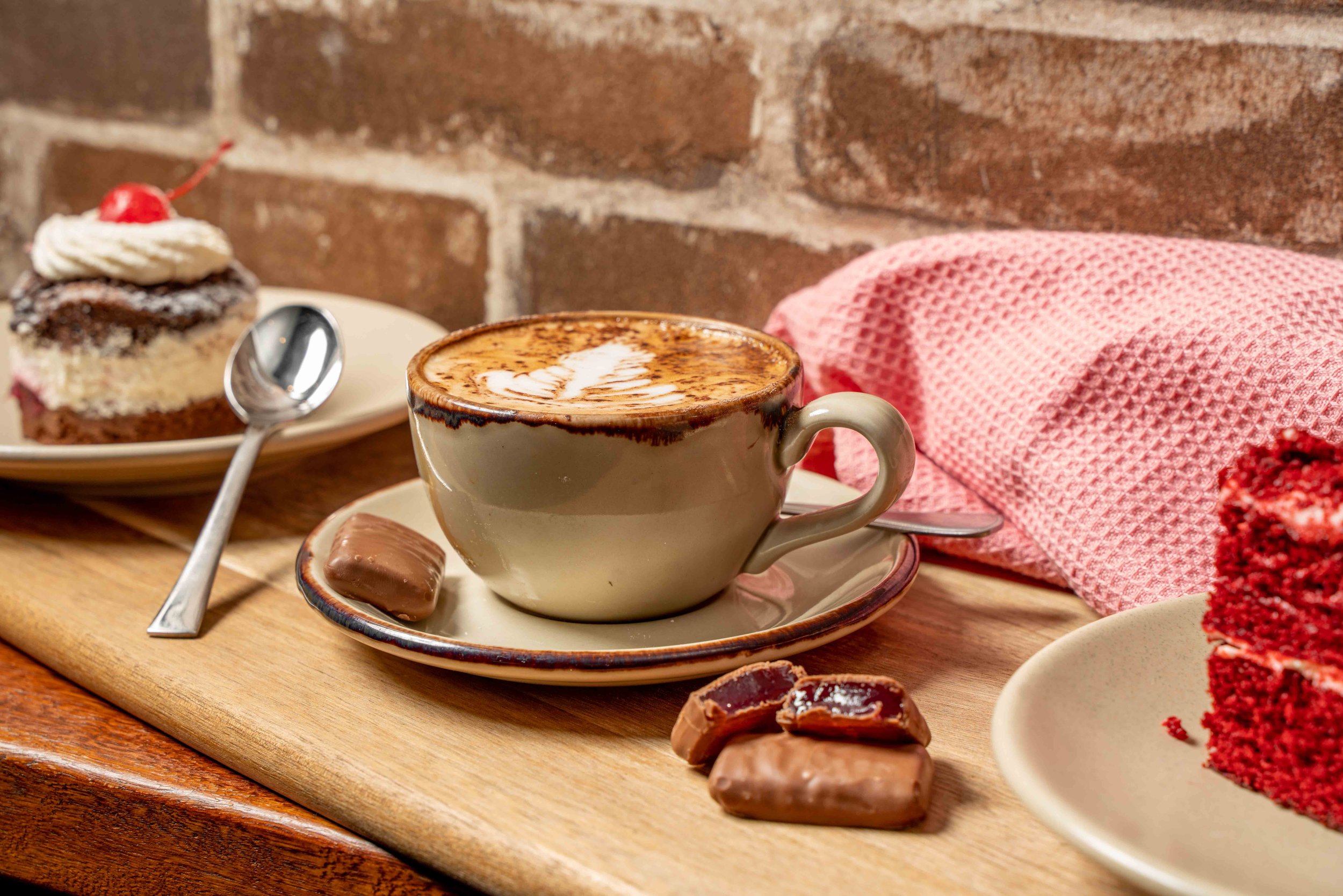 A cup of cappuccino with latte art on top, a partially unwrapped chocolate candy on a saucer, a slice of layered cake with whipped cream and a cherry on top on a plate, and broken pieces of chocolate on a wooden table against a brick wall.