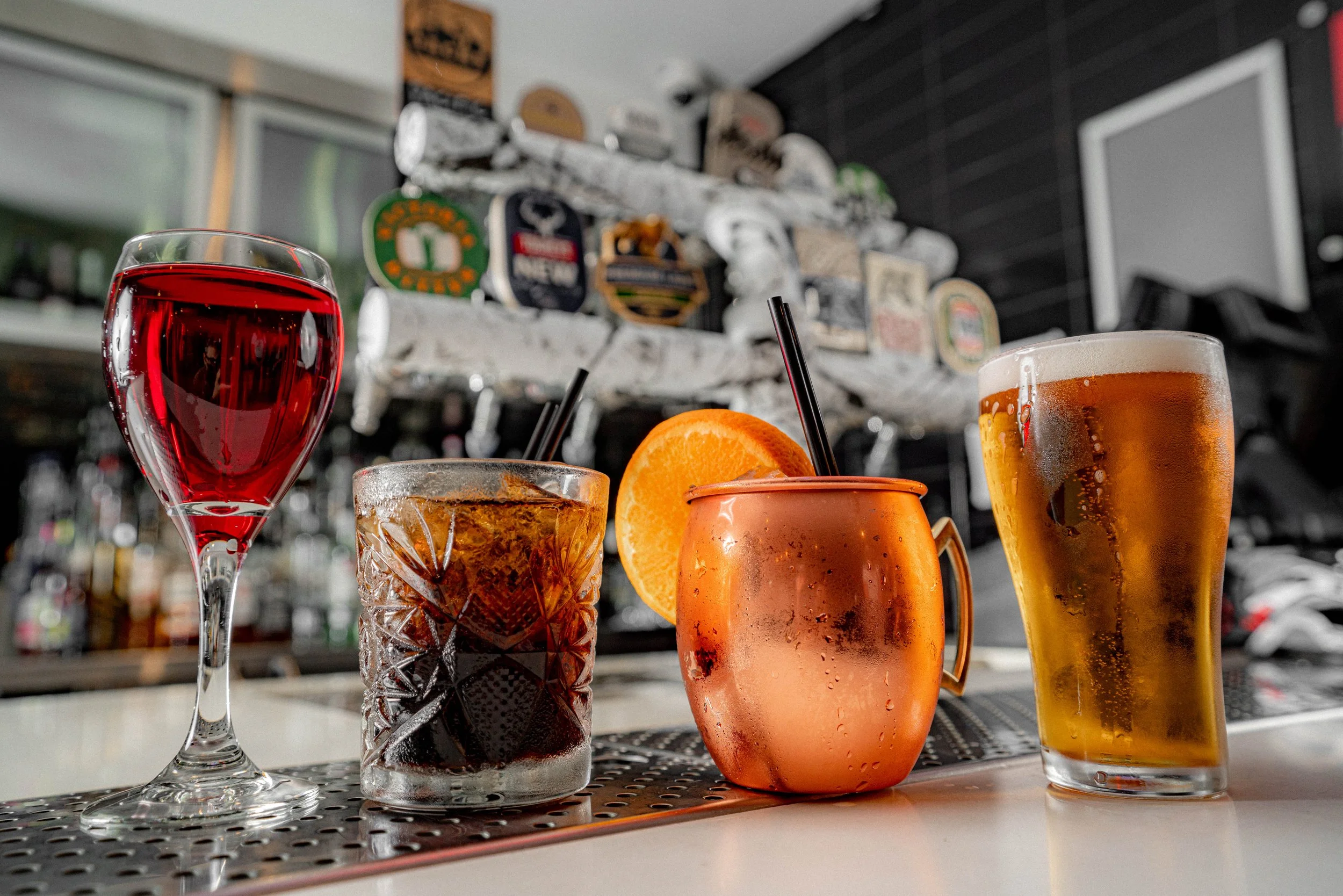 A variety of colorful cocktails with an orange garnish and black straws on a bar counter, with beer taps and beer signs in the background.