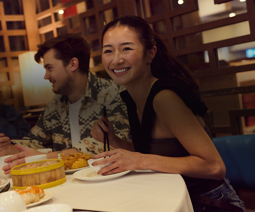 A group of young adults enjoying a meal at a restaurant, smiling and talking, with various dishes on the table.