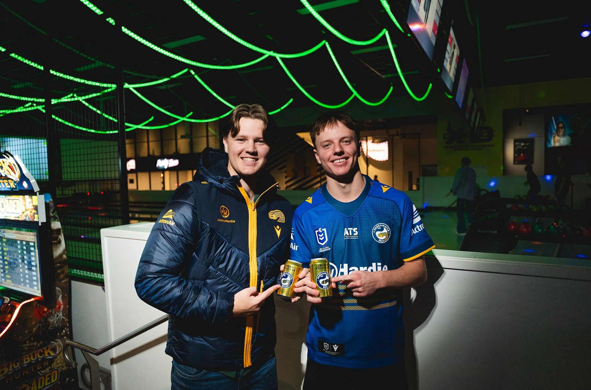 Two young men in a gaming arcade, smiling and holding cans of beverages, one wearing a dark jacket and the other in a blue sports jersey, under bright green neon lights.