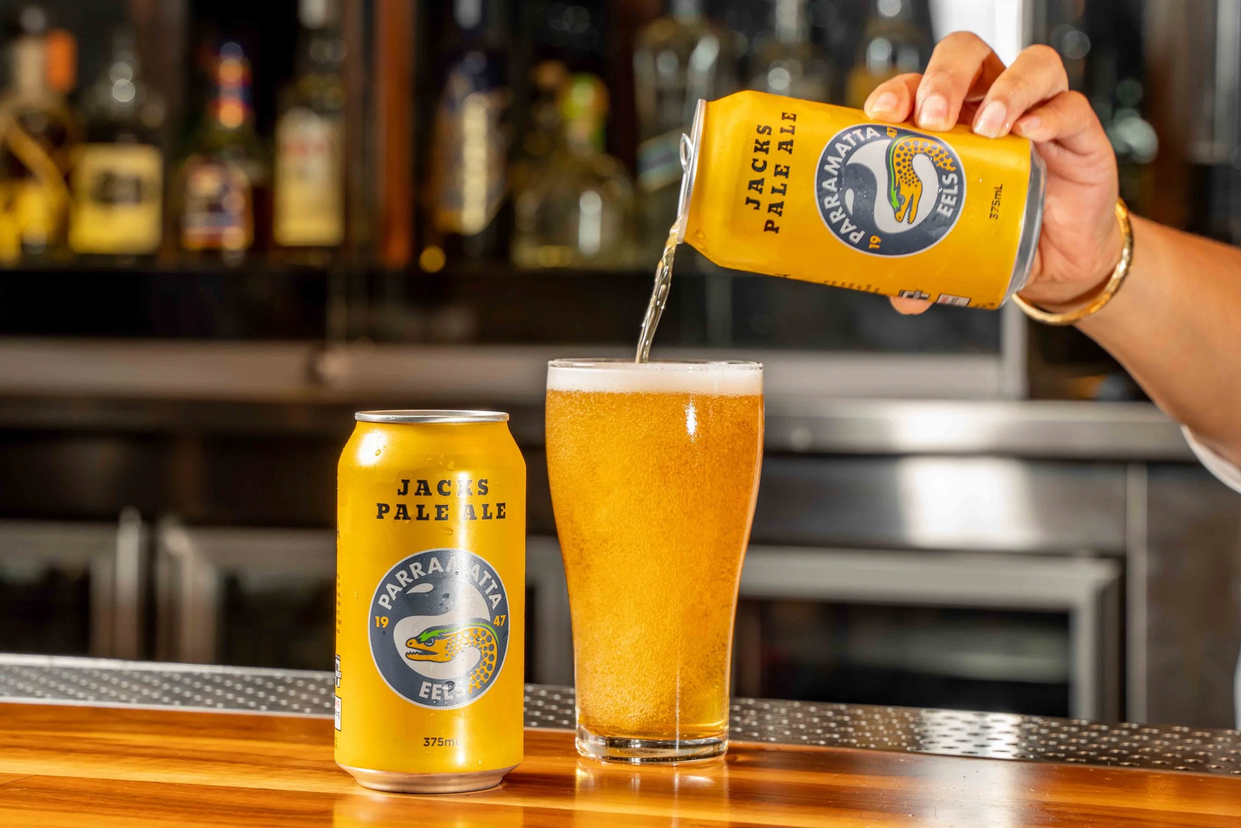 A hand is pouring beer from a yellow beer can labeled 'Jack's Pale Ale' into a glass filled with beer. There is an unopened beer can on a wooden surface in the foreground, and a blurred bar background with multiple bottles.