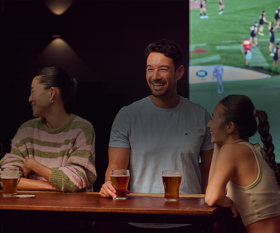 Group of people socializing at a bar with drinks, smiling and engaging in conversation, with a television displaying a sports game in the background.
