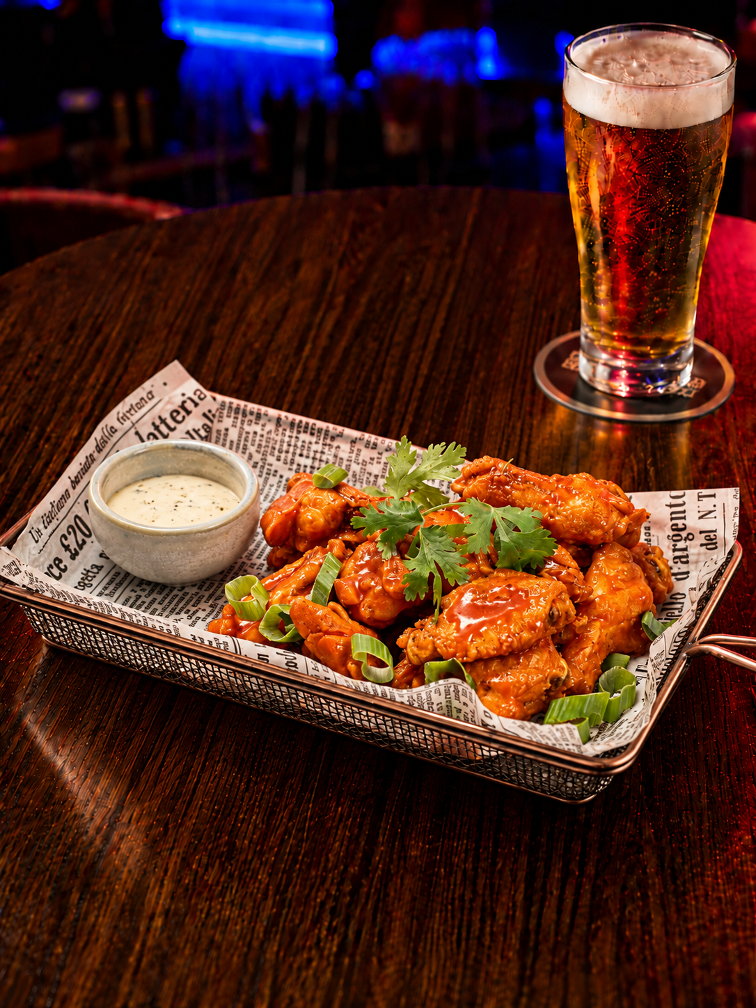 Basket of buffalo chicken wings with celery sticks, ranch dressing, a glass of beer, and a small bowl of dipping sauce on a dark wooden table in a sports bar.