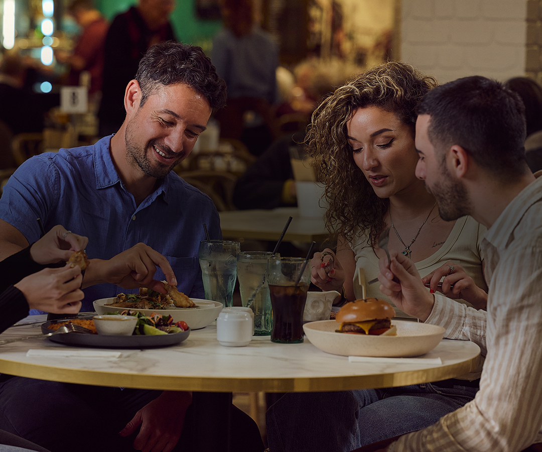 A group of five diverse young adults sitting around a table in a restaurant, enjoying a meal together and smiling. The table has various dishes and drinks, including a burger, salad, and beverages. The background shows other diners and a cozy, warmly lit atmosphere.
