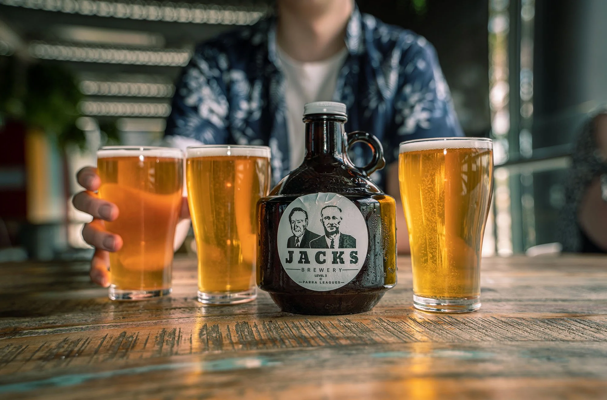 Four glasses of beer on a wooden table with a dark glass bottle labeled 'Jacks Brewery' in the center. Someone is reaching toward the glasses, which are filled with amber-colored beer. A person wearing a patterned blue shirt is sitting behind the table, but only their upper torso and head are partially visible.