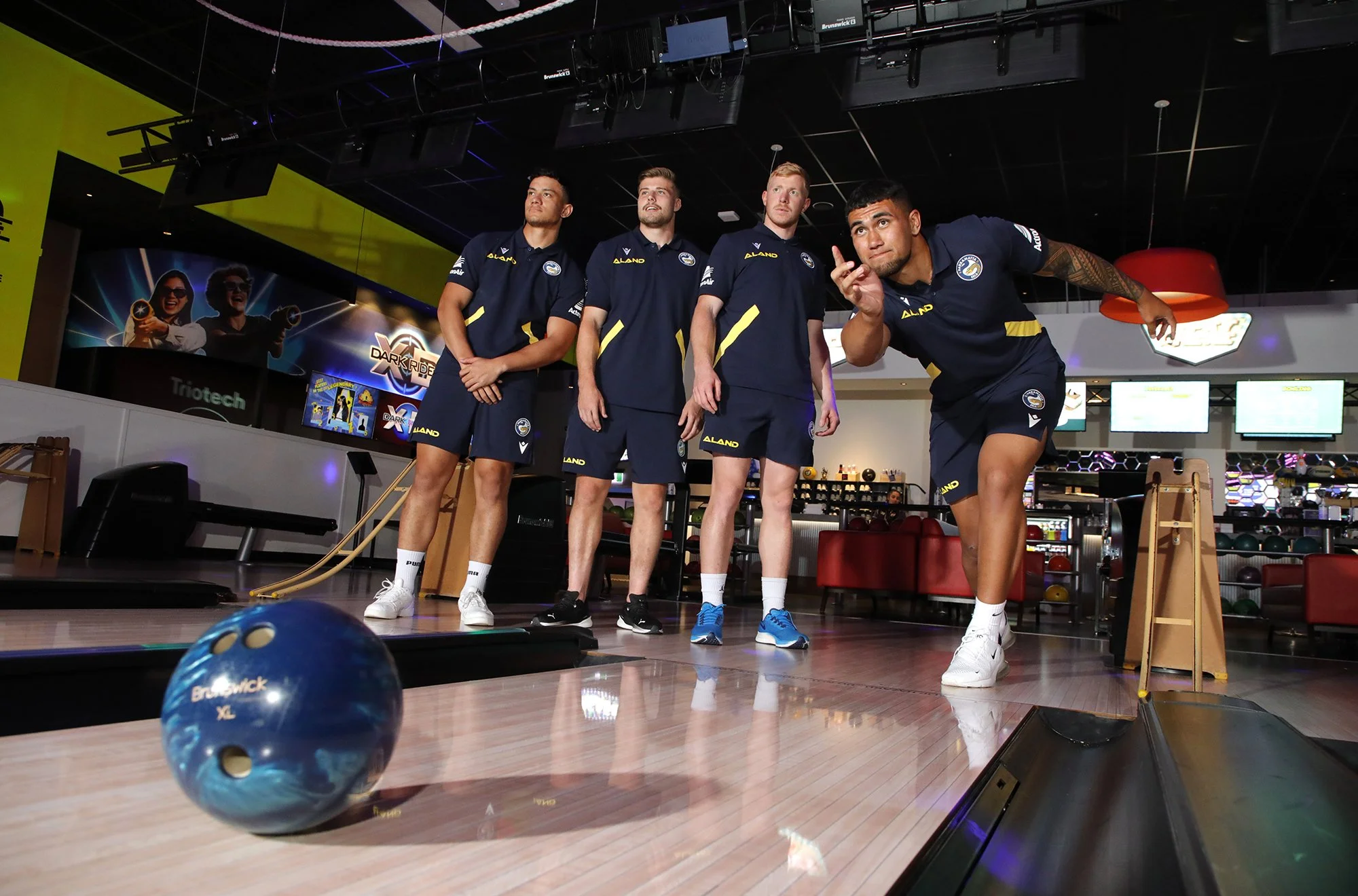 Four men in navy blue sports uniforms standing on a bowling alley lane, one preparing to roll a blue bowling ball. The background features arcade and entertainment signs, including one for Dark Ride, and arcade machines.