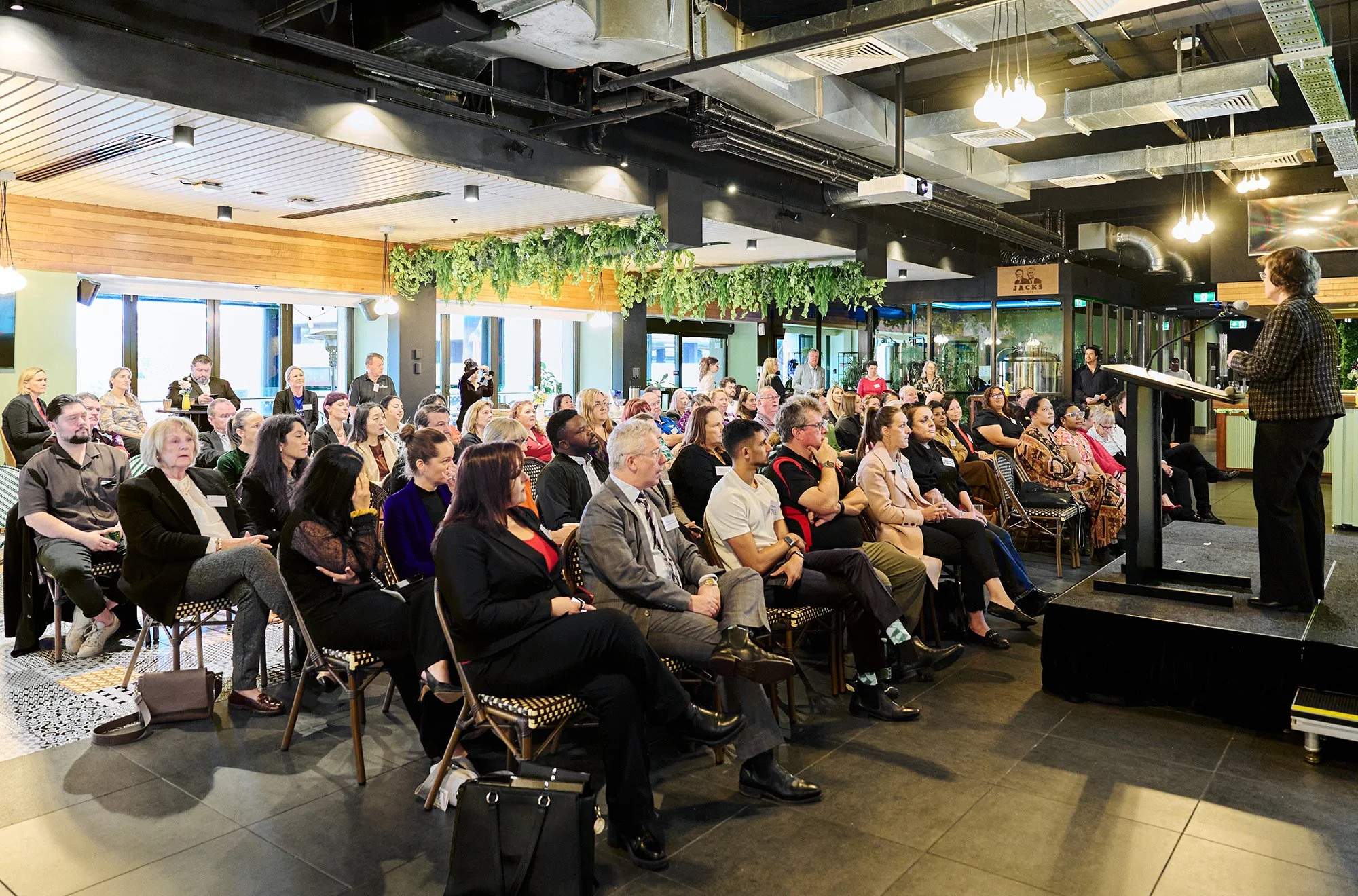 A large group of people seated in a conference room listening to a woman speaking at a podium. The room has modern decor with hanging greenery, wood accents, and industrial-style ceilings.