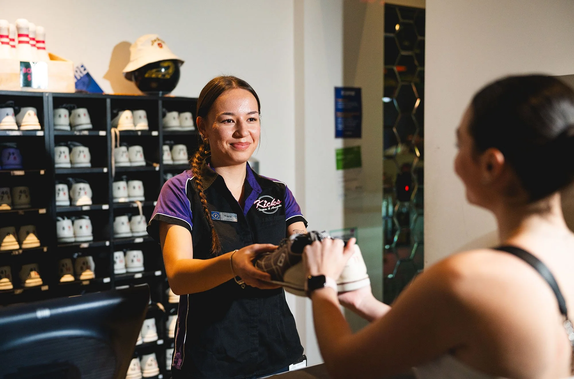 A female employee at a retail store handing a white sneaker to a customer. The background shows shelves filled with various shoes and a display with a hat on it.