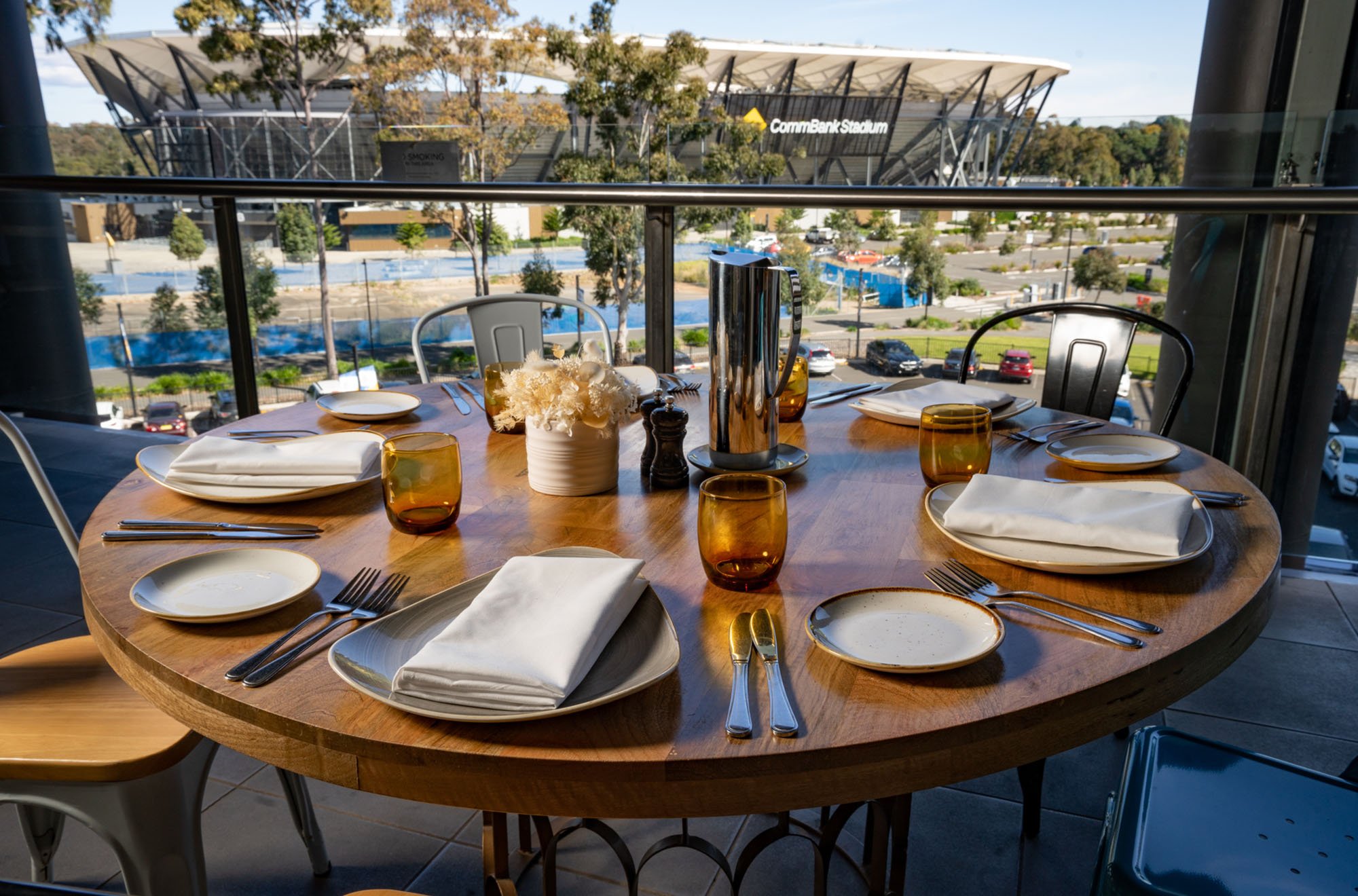 Round wooden dining table set for six with white napkins, clear amber glasses, white plates, silver cutlery, and a white flower centerpiece at an outdoor restaurant. The background shows a parking lot, trees, and a building labeled 'CommBank Stadium'.