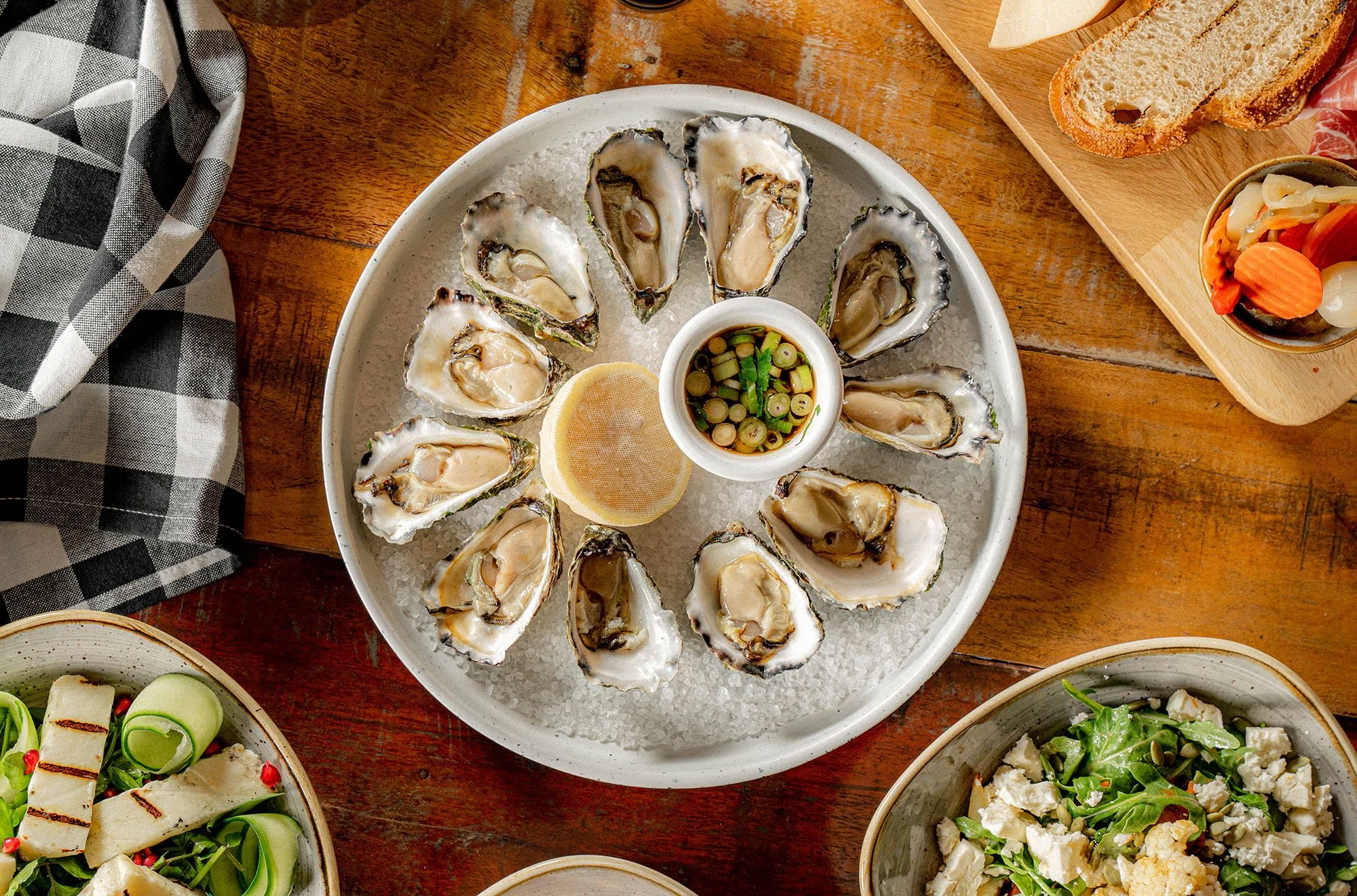 A plate of fresh oysters on ice with a lemon wedge and dipping sauce in a small bowl at the center, surrounded by various dishes and wooden table surfaces.