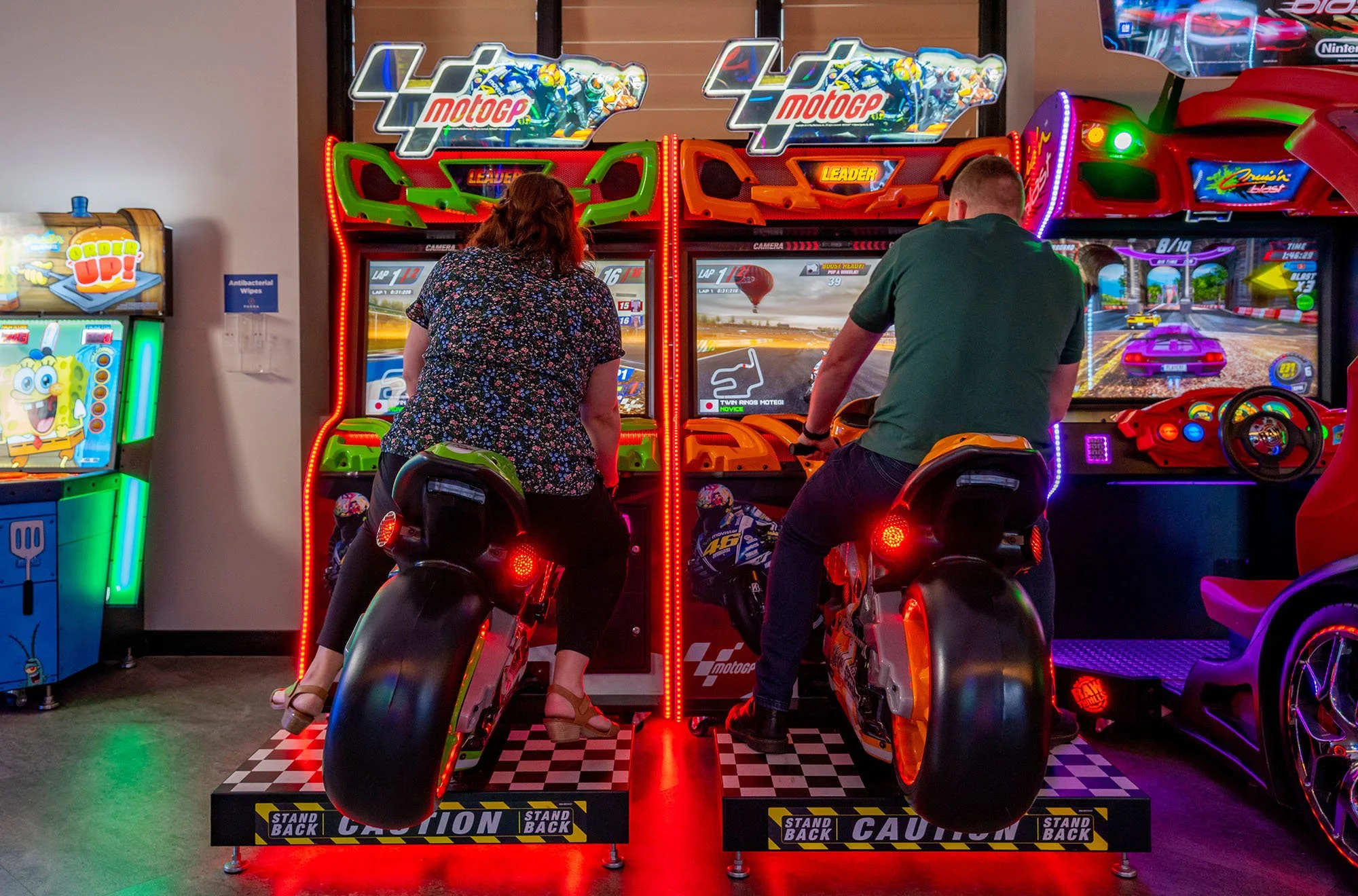 Two children riding on motorbike arcade game in an arcade. The game features a racing theme with a motorcycle cockpit style. Bright, colorful lights and digital screens display racing scenes.