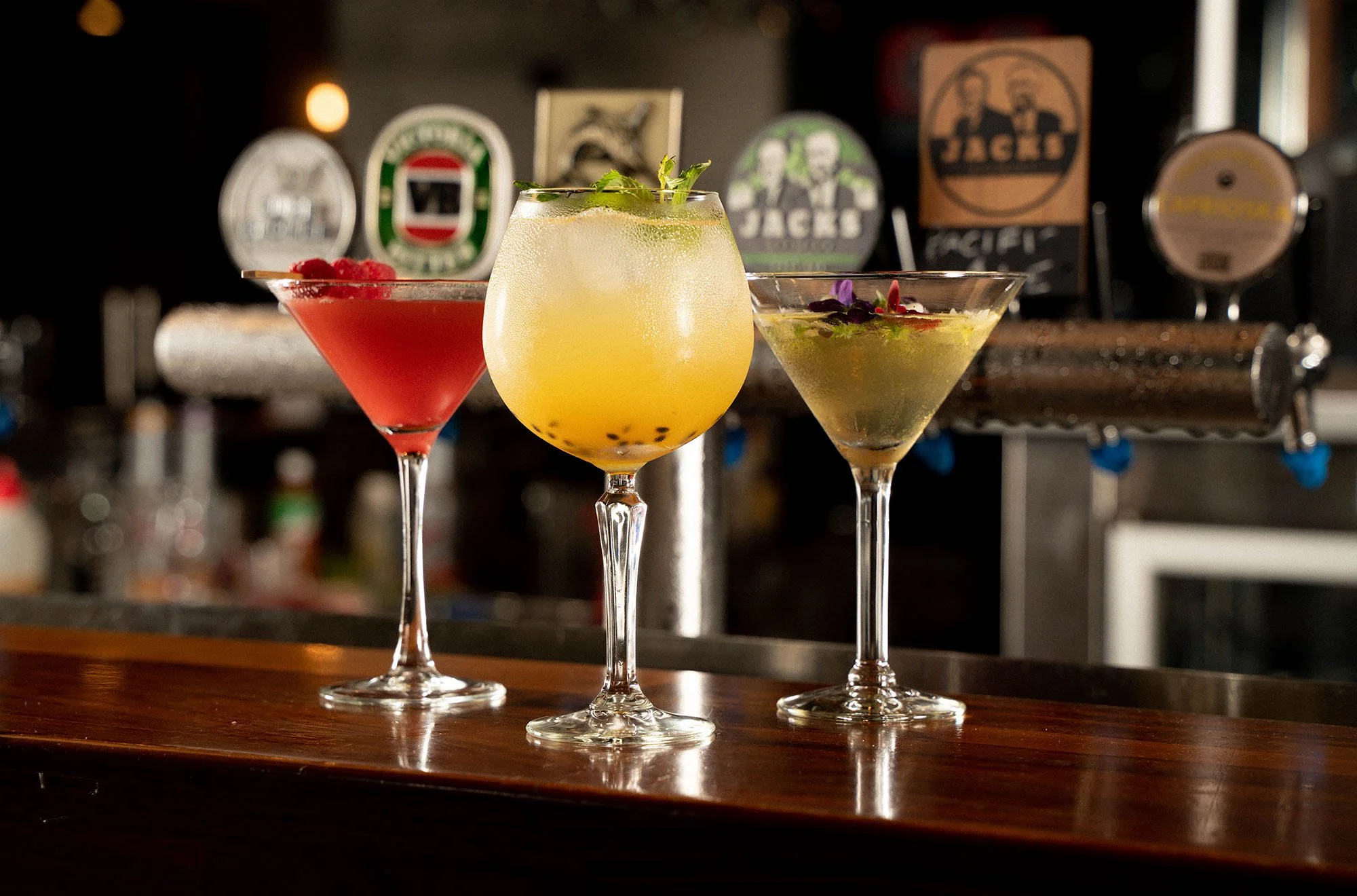 Three cocktails in various glasses placed on a bar counter, with a blurred background showing bar decorations and taps.