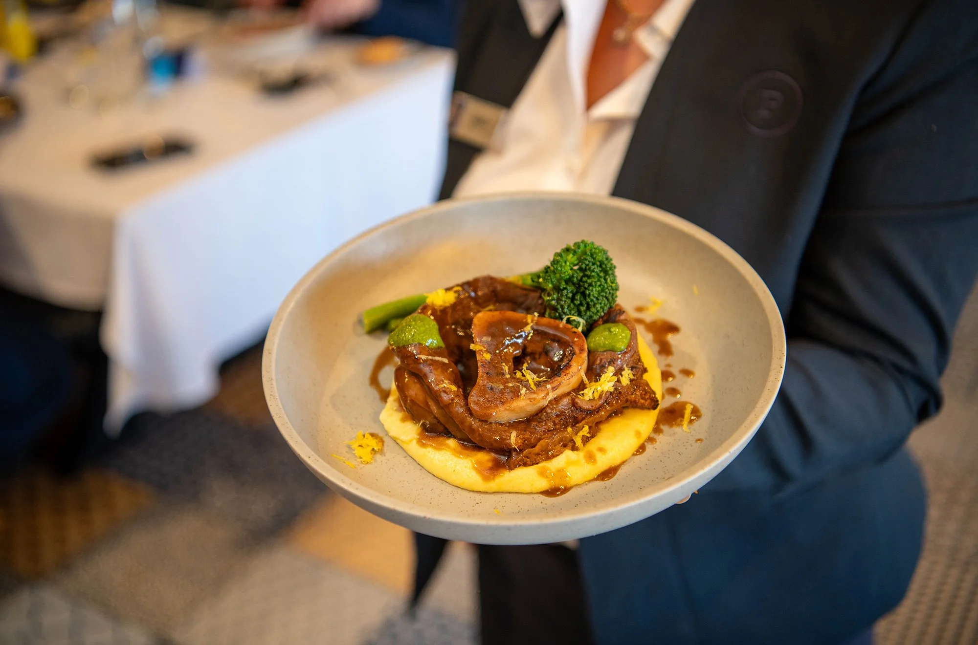 A person in a dark suit holding a white bowl containing meat, broccoli, and mashed potatoes with gravy.