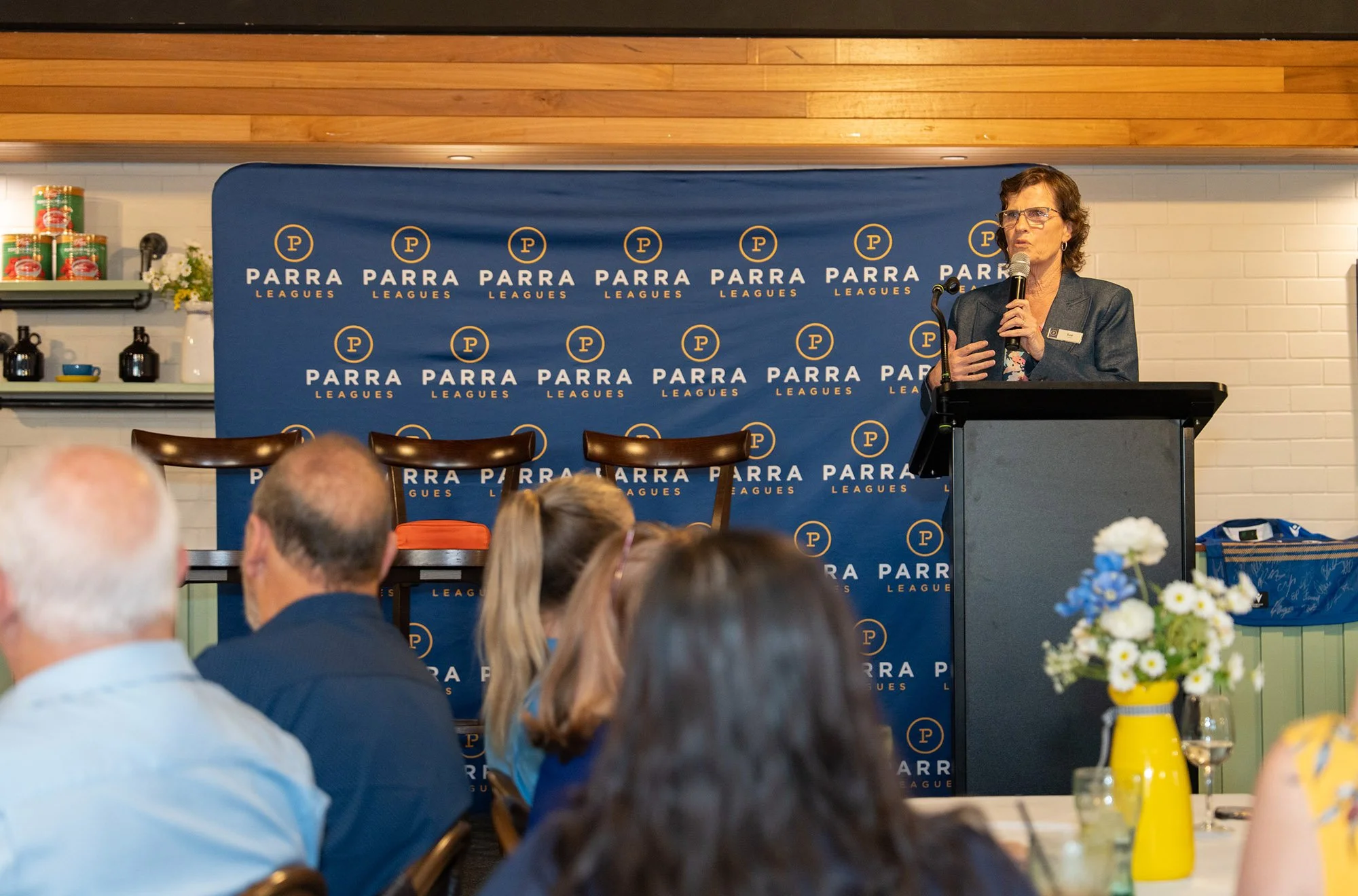 A woman speaking at a podium during an event with a blue backdrop branded with 'Parra Leagues'. Audience members are seated in front of her, with a table decorated with a yellow vase and flowers visible in the foreground.