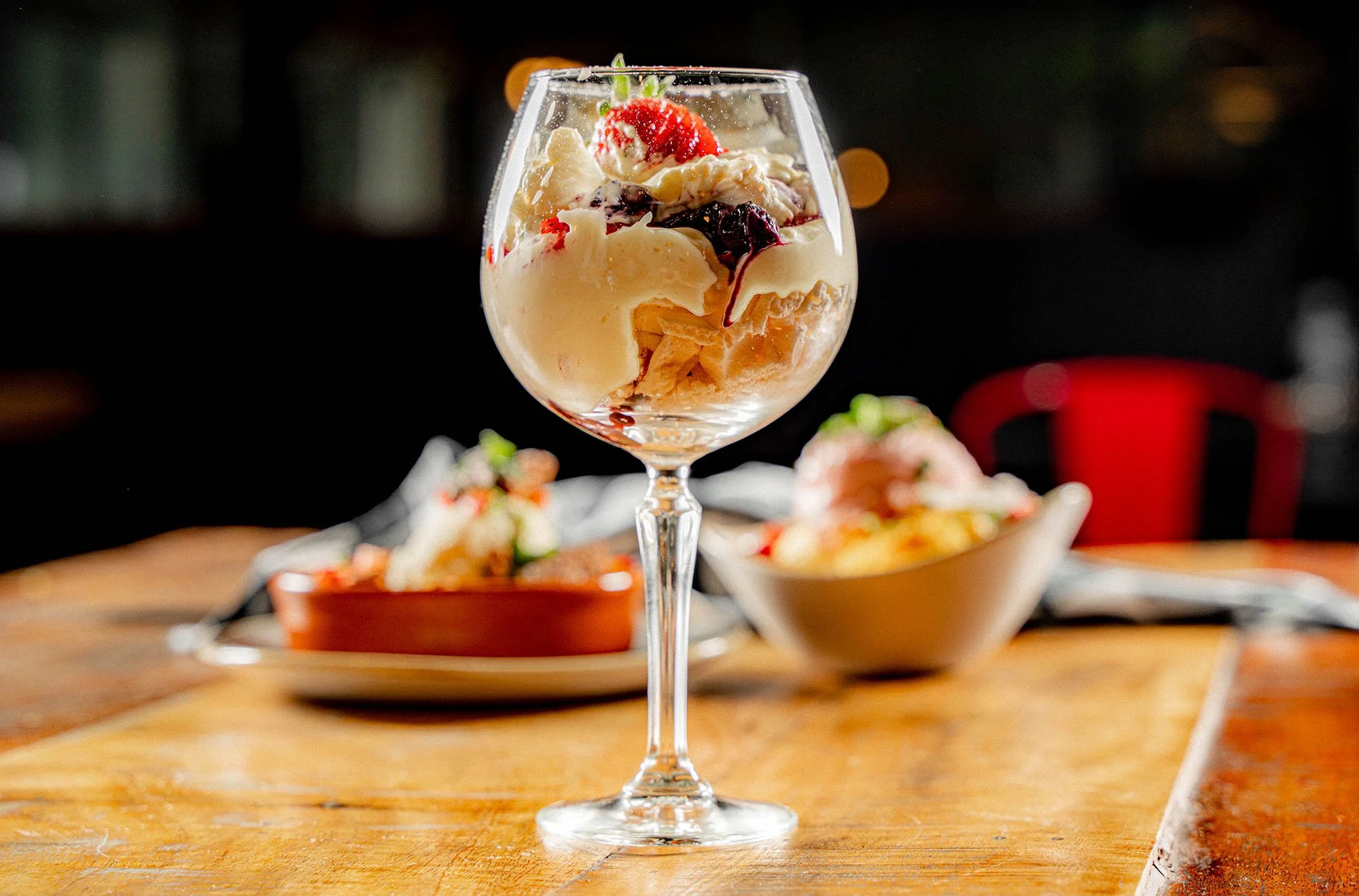 A glass dessert cup filled with vanilla ice cream, topped with strawberries and berry syrup, on a wooden table, with bowls of dessert in the background.