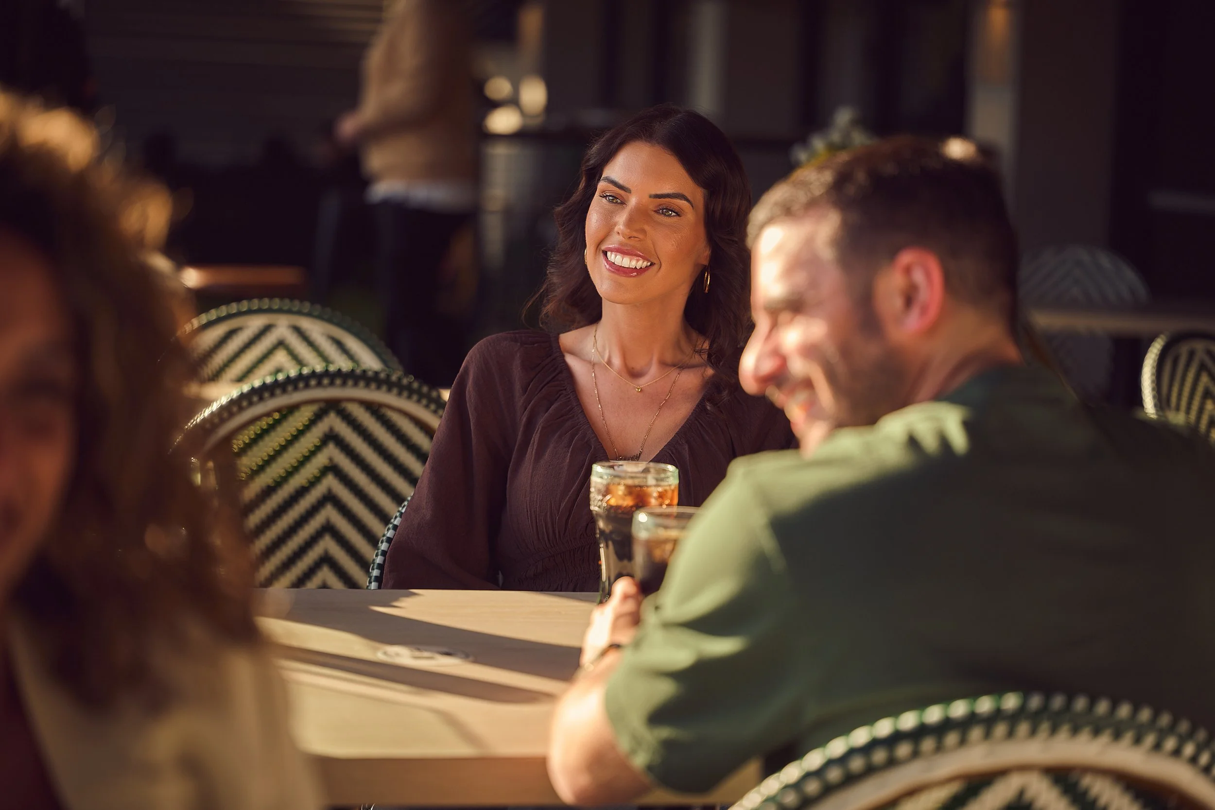 A group of people socializing at a bar or restaurant, focusing on a woman with dark hair smiling and looking at someone outside the frame. Others are holding drinks and talking.