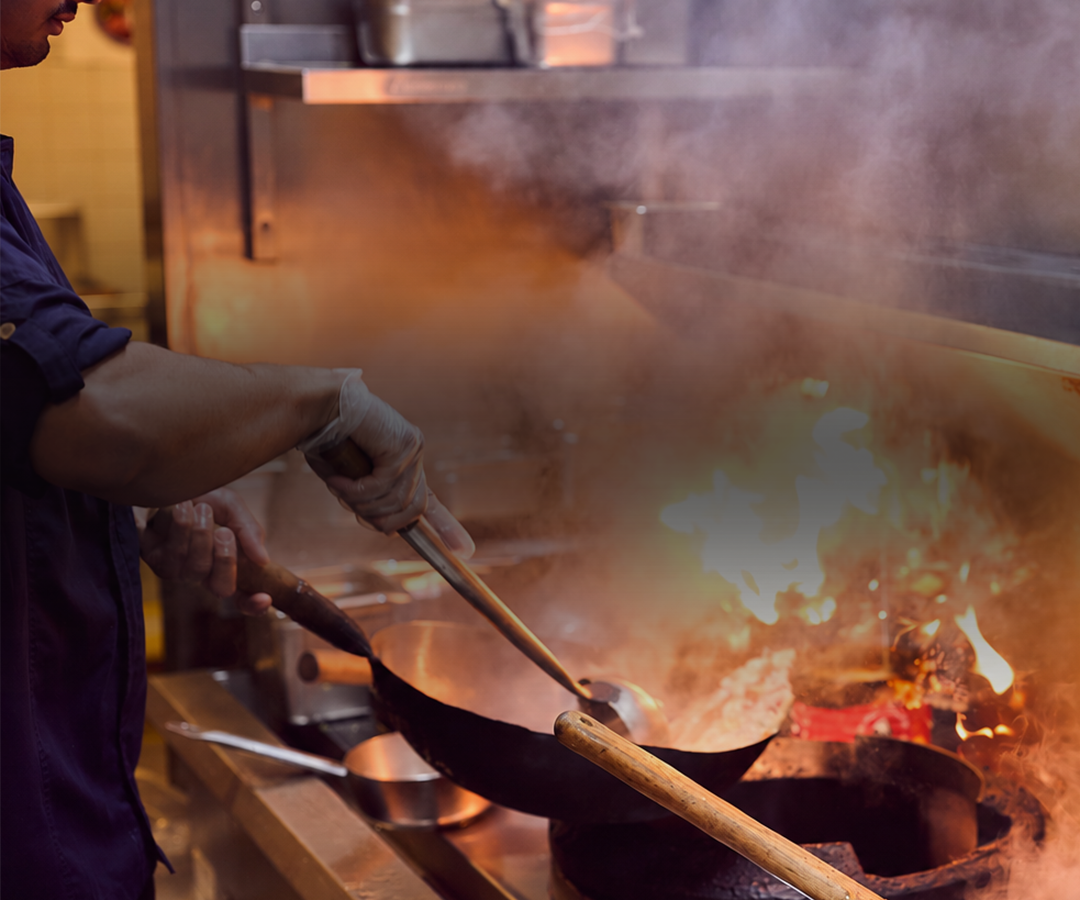 Chef cooking with a flame in a professional kitchen, stirring a pan on a stove.