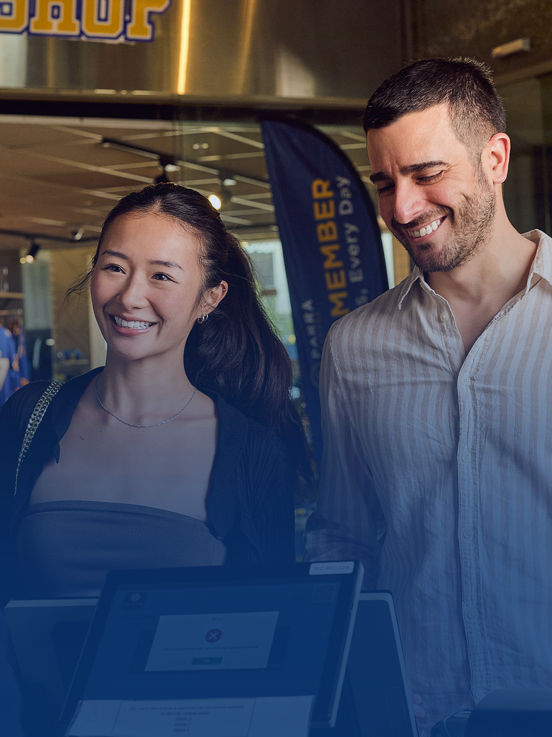 Two women and a man smiling at each other in a store, with a cashier and a checkout screen visible in the foreground. The store has a sign that says 'CLUB SHOP' and clothing racks in the background.