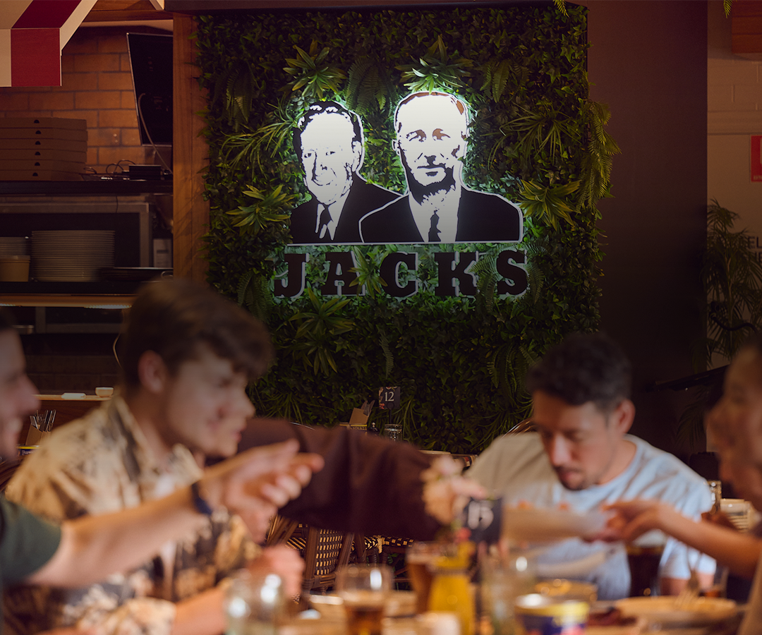 A lively restaurant scene with multiple people dining and talking at a table, featuring a green leafy wall with illuminated black and white portraits and the text 'JACKS.'