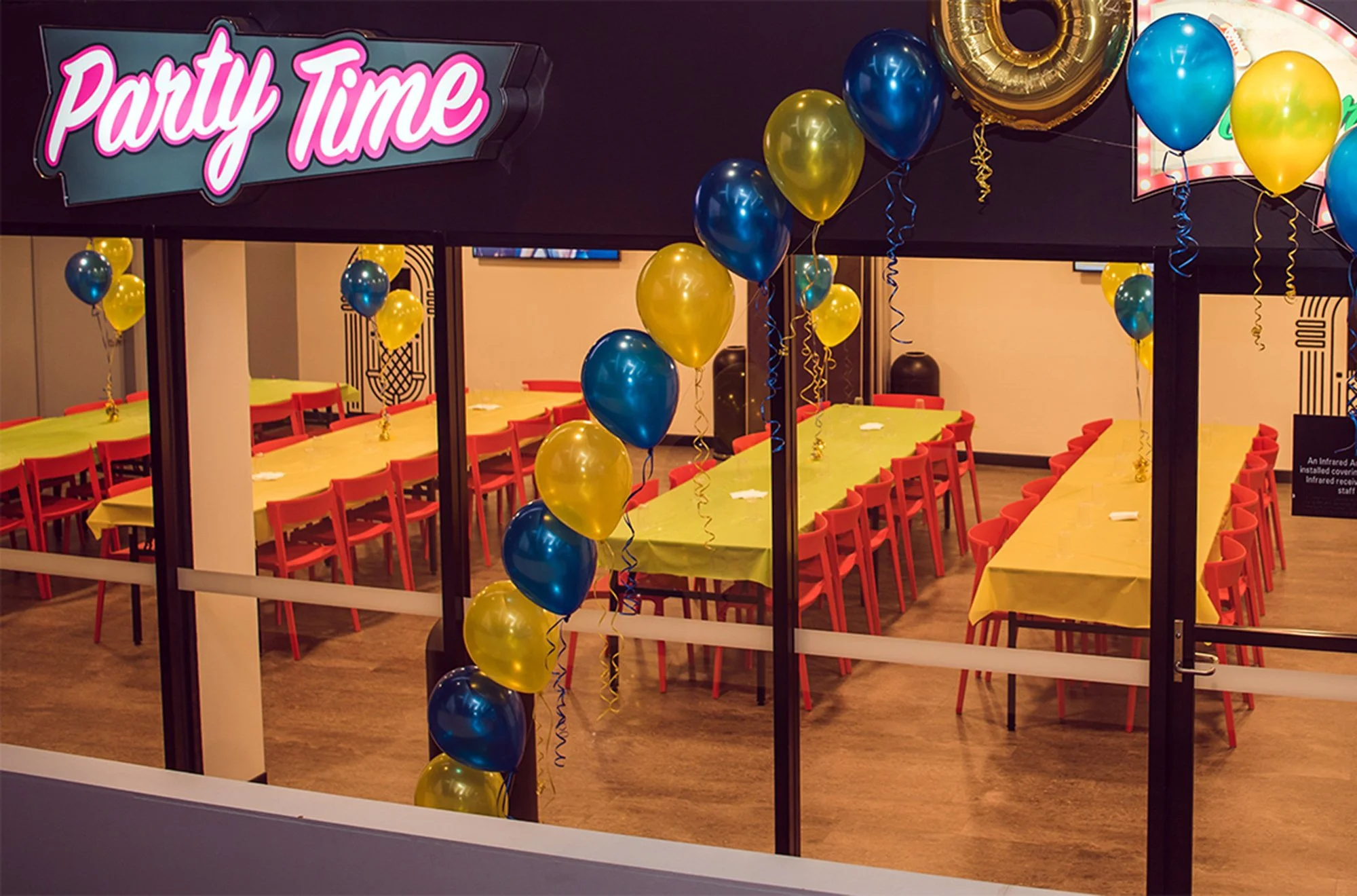 Colorful party room decorated with yellow and blue balloons, with a sign that reads 'Party Time,' featuring long tables covered with yellow tablecloths and red chairs for guests.