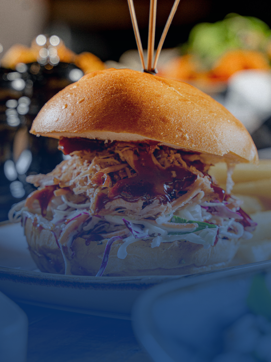 A close-up of a pulled pork slider sandwich with shredded pork, coleslaw, and barbecue sauce in a dinner roll, with a plastic heart-shaped toothpick on top.
