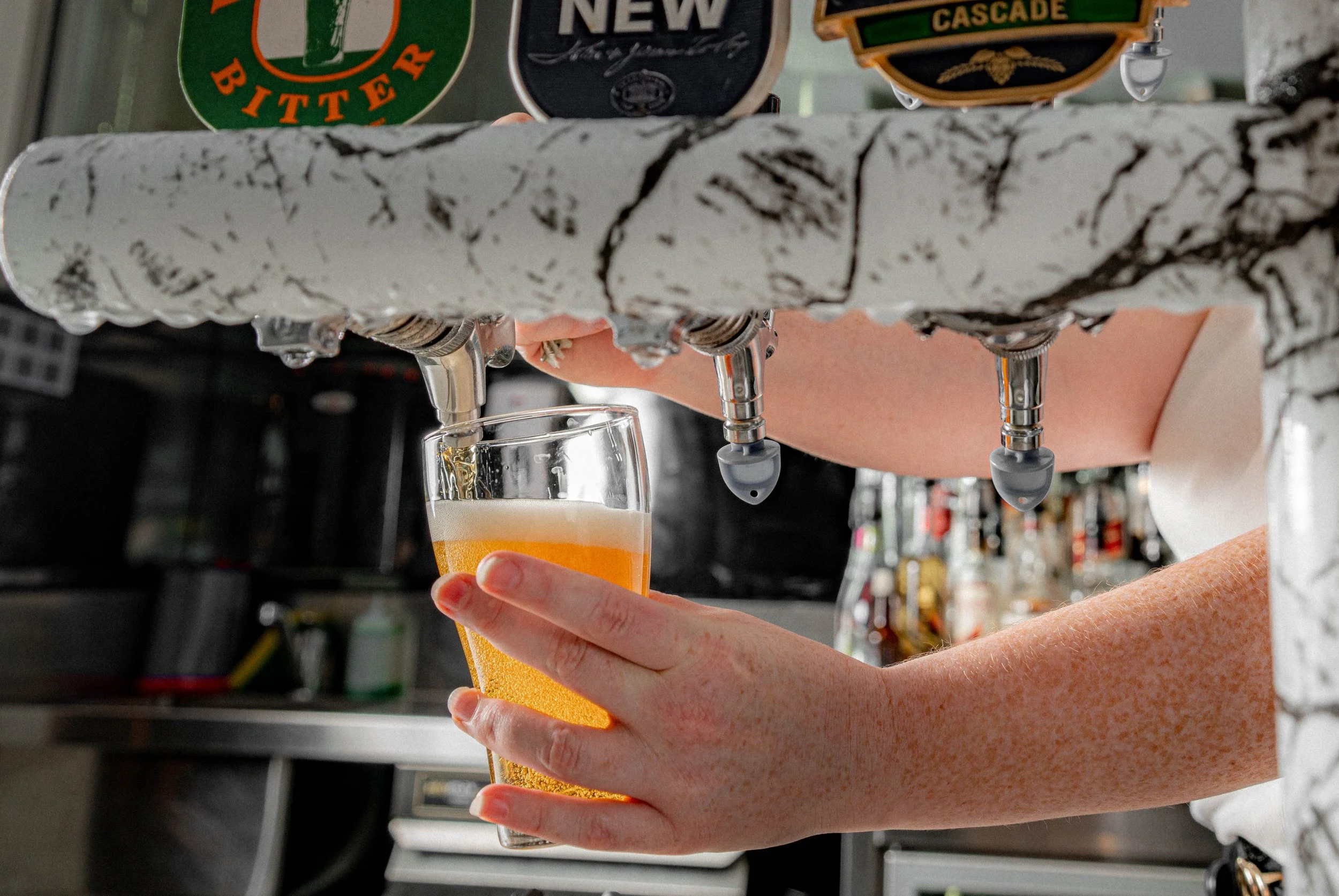 Person pouring draft beer into a glass using a tap from a beer dispenser with various beer signs in the background.