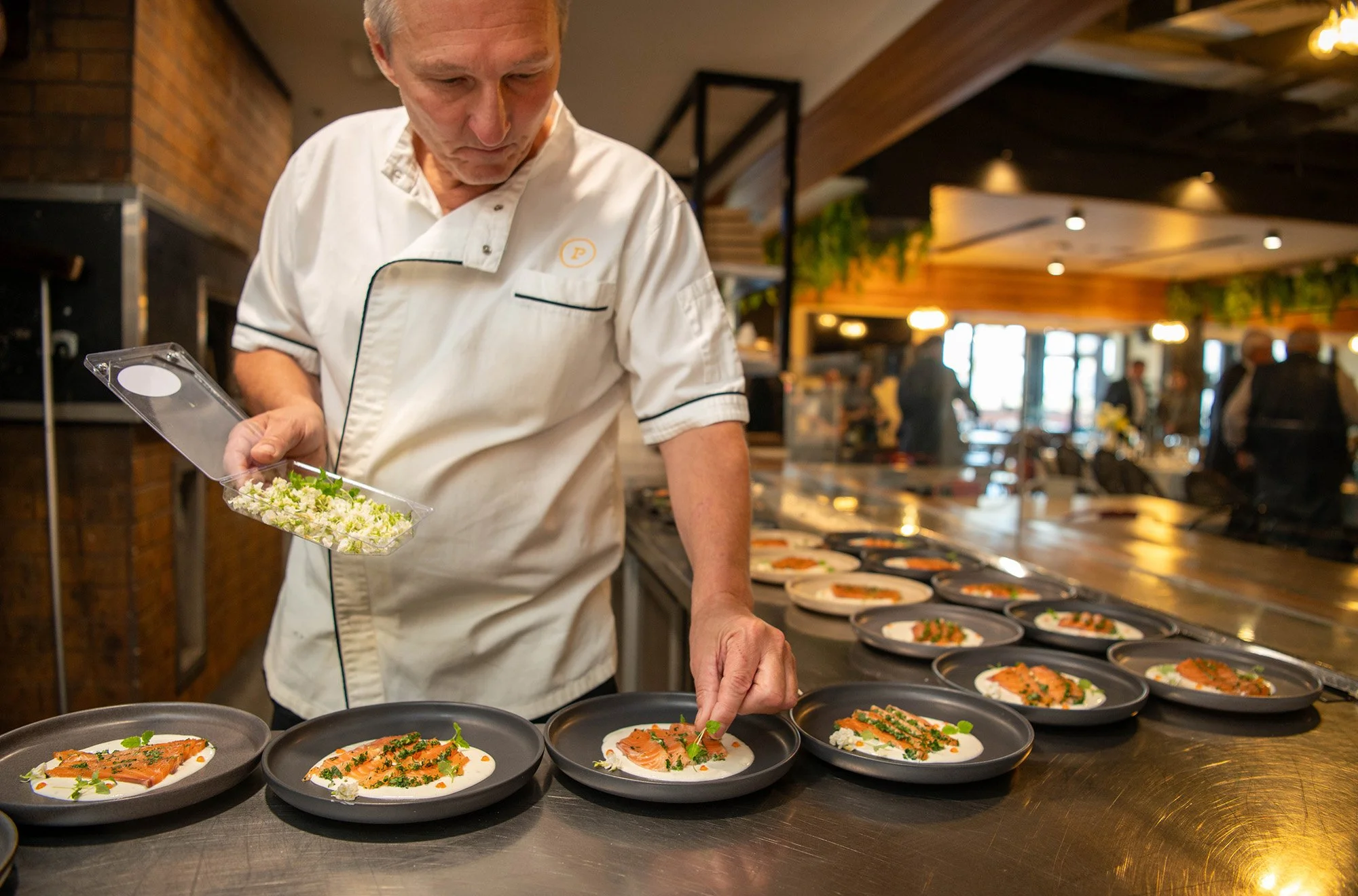 A male chef in a white uniform garnishes plates of salmon on a restaurant serving counter.