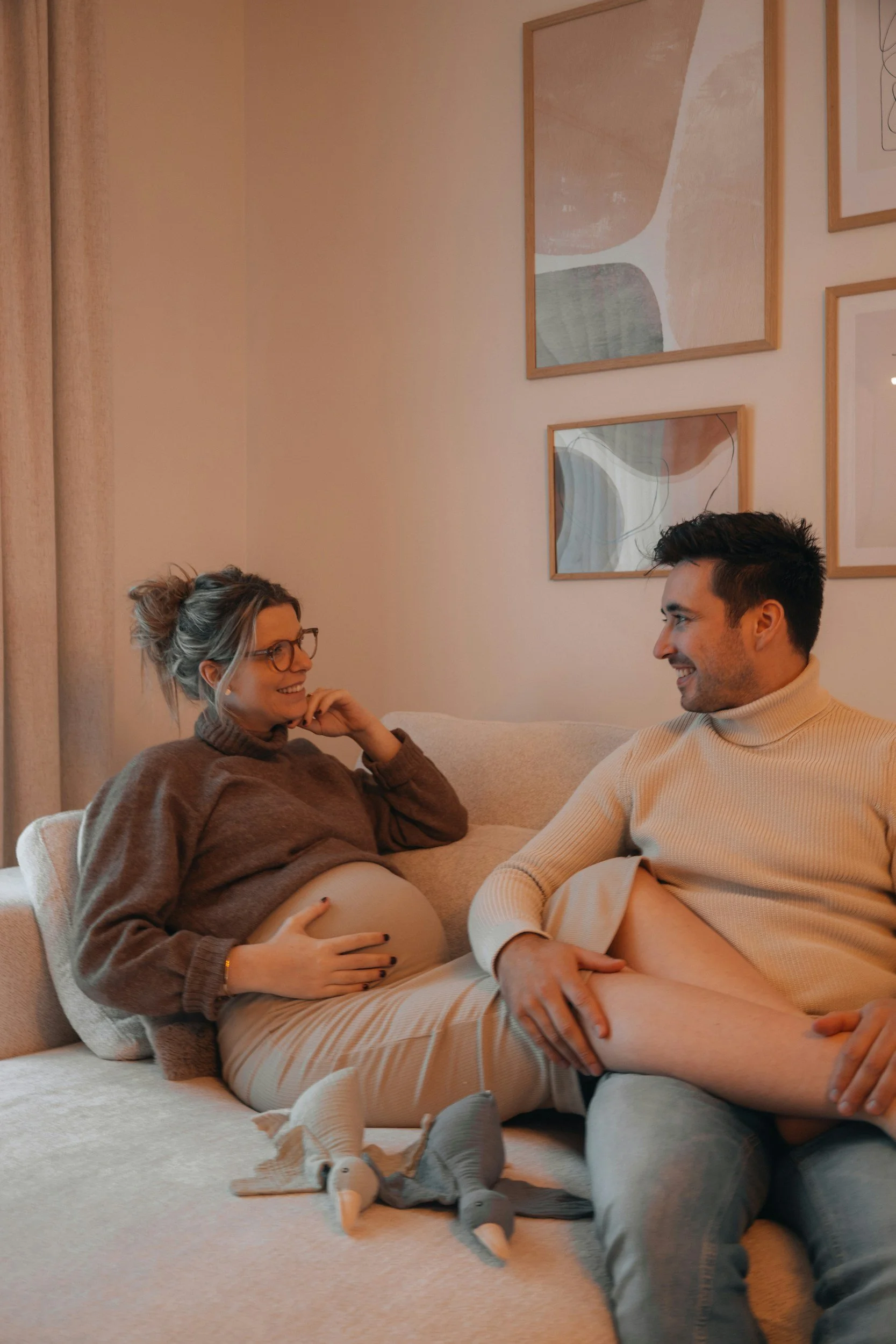 A couple sitting on a beige couch, smiling and engaging in conversation. The woman is pregnant, wearing a brown sweater and cream pants, holding her belly with one hand. The man is wearing a cream turtleneck and appears to be talking to her. The wall behind them is decorated with framed abstract artwork and beige curtains.