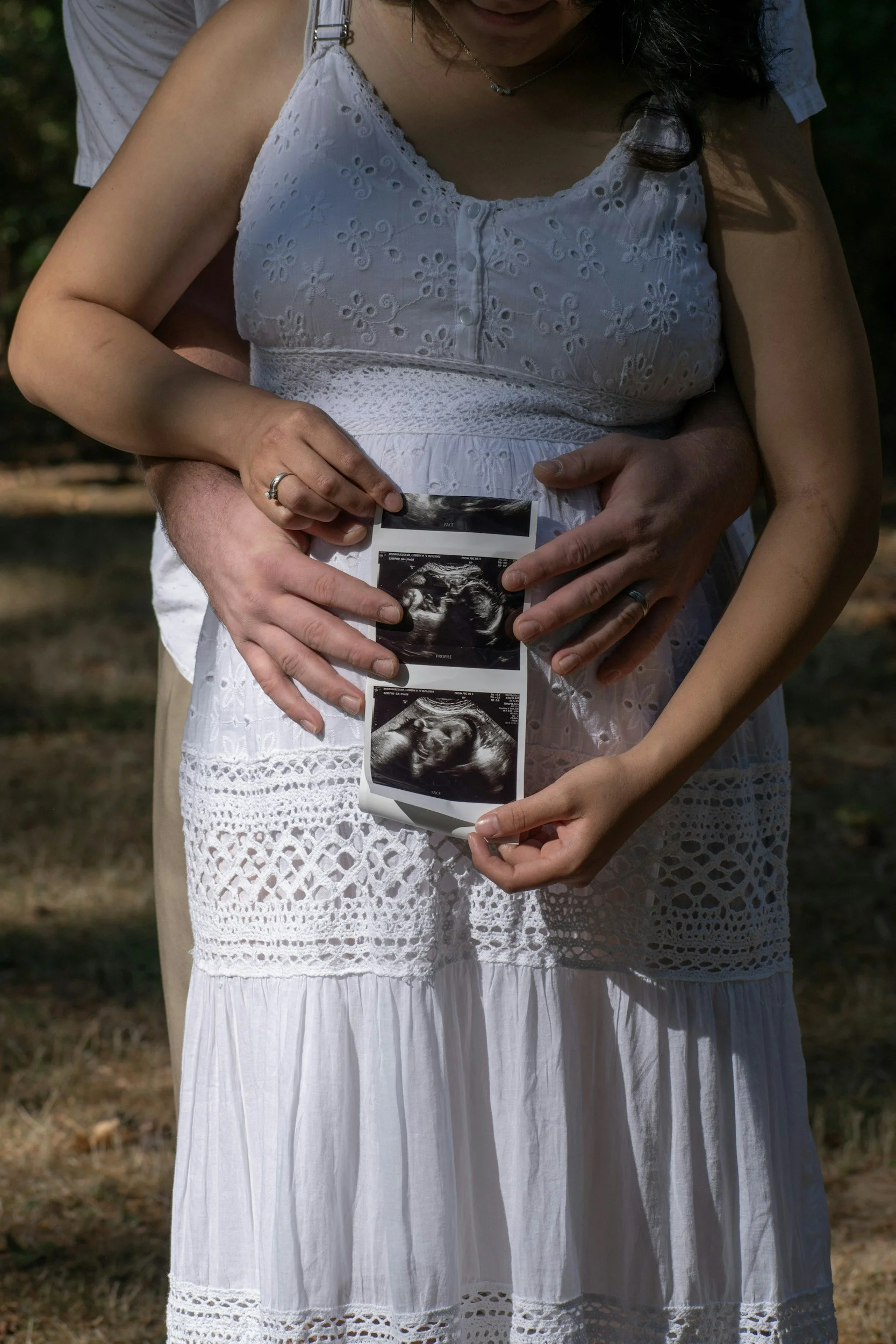 A pregnant woman in a white dress holding ultrasound images of her baby, with a man behind her embracing her.