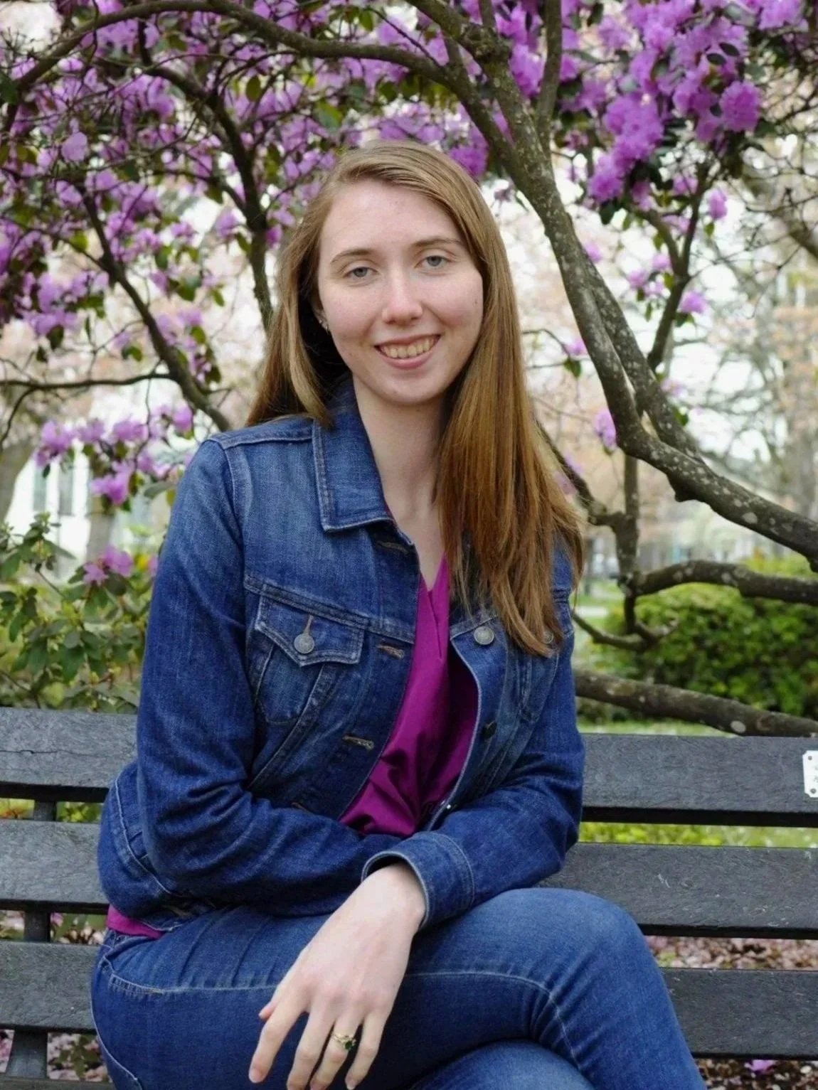 A young woman with long red hair, wearing a denim jacket and purple top, sitting on a park bench surrounded by blooming pink and purple flowers on trees.