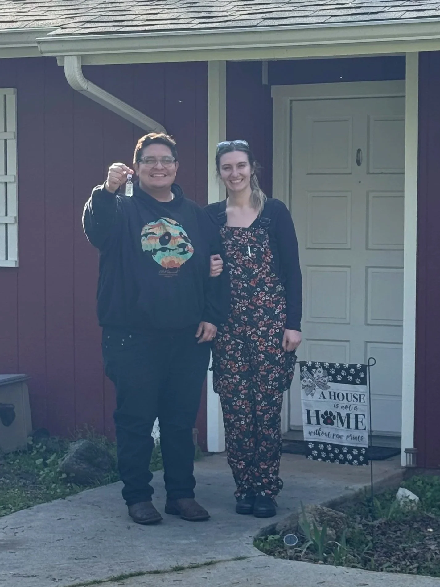 A man and woman standing in front of a house with a sign that reads "A house is not a home without our paws." The man is holding a small object, possibly a key, and they are smiling at the camera. The house has a purple exterior with a closed garage door.