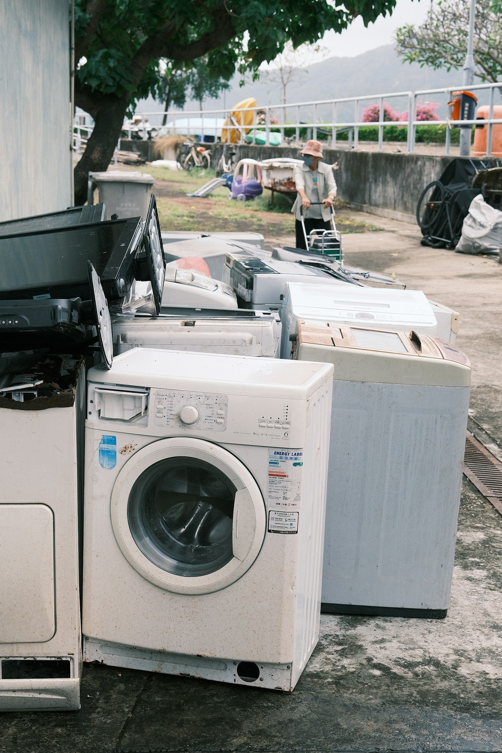 Several discarded washing machines and appliances outdoors near a wall, with a person pushing a cart and trees in the background.