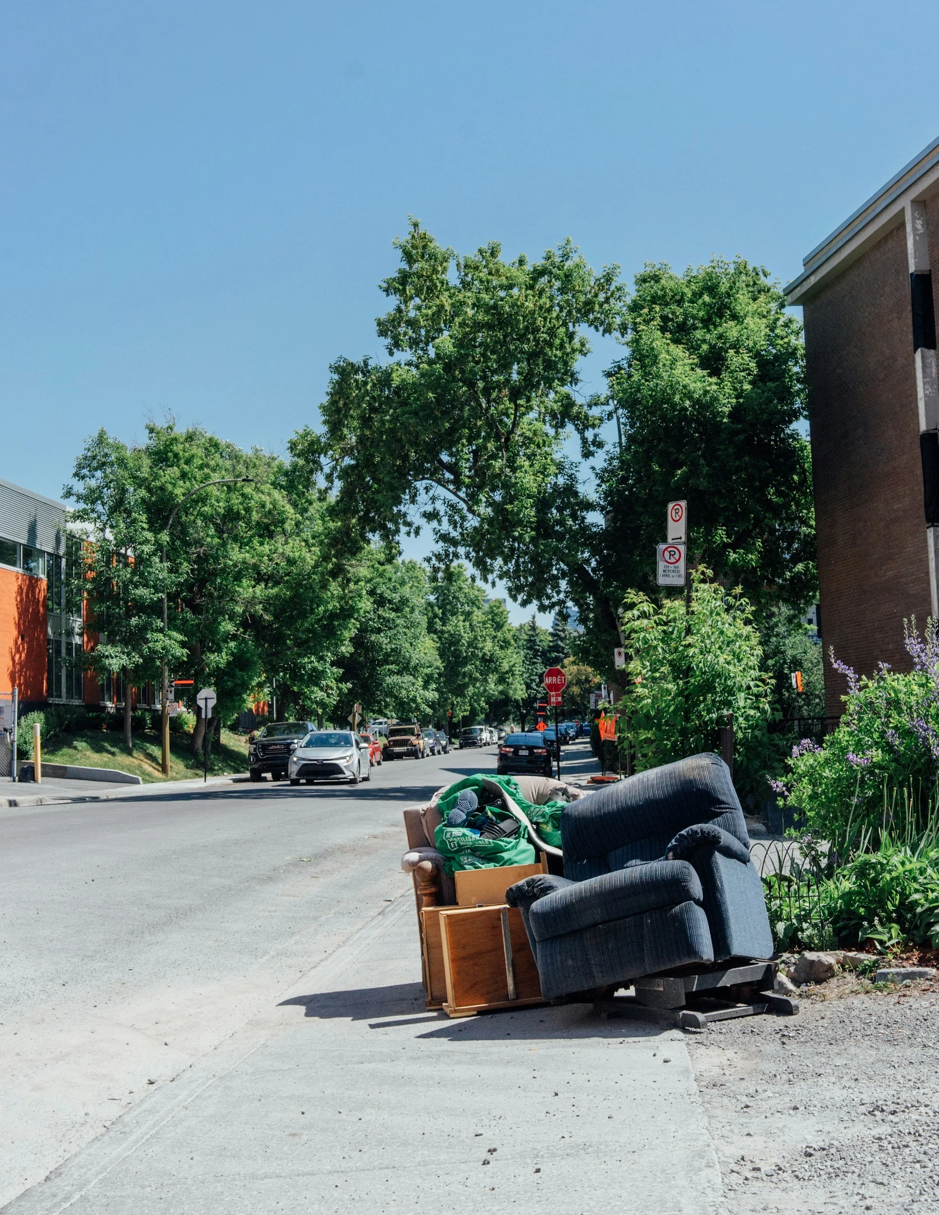 A street scene with trees, parked cars, and buildings. In the foreground, there are discarded furniture items including a chair and a sofa with blankets and household items on them.