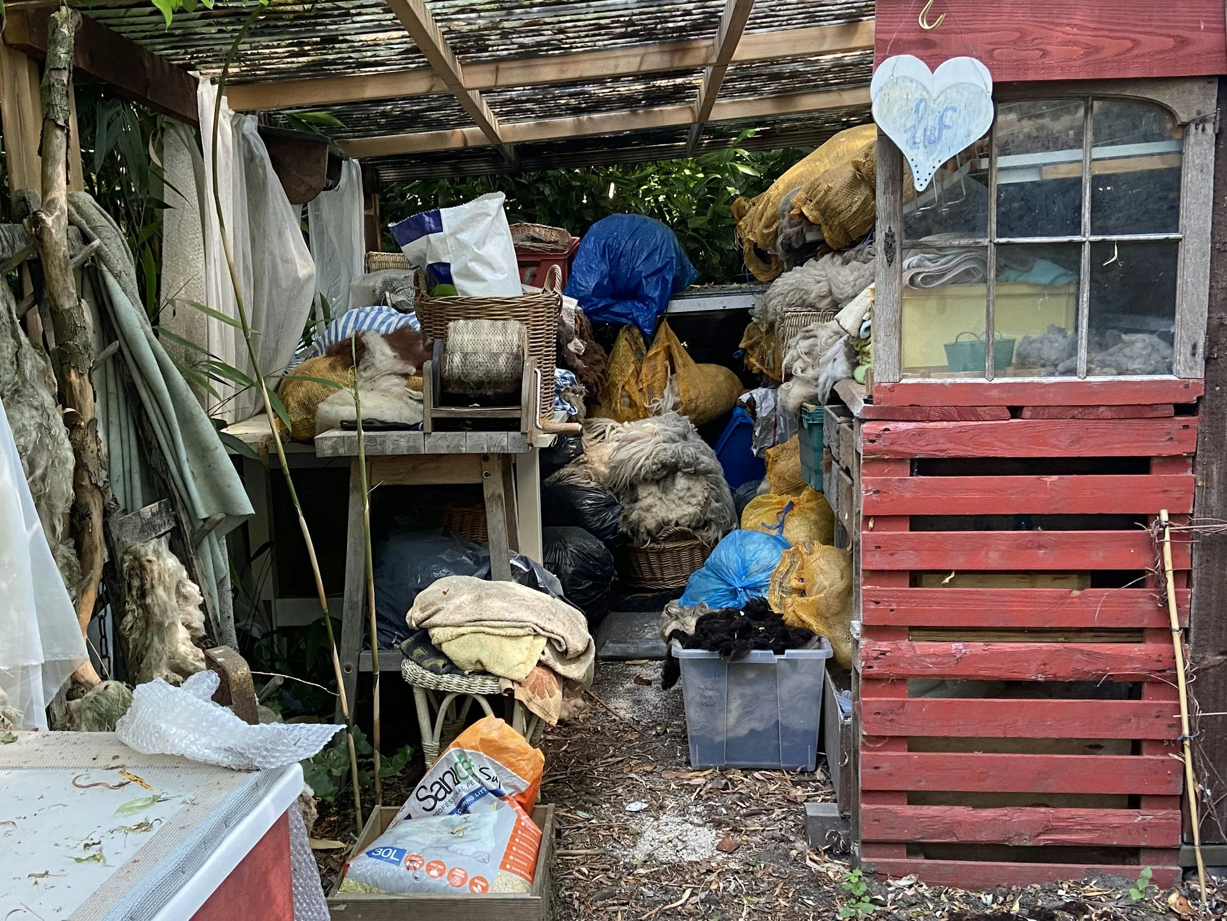 Covered outdoor shed filled with miscellaneous items including bags, fabric, and boxes, with a red wooden fence on the right and greenery in the background.