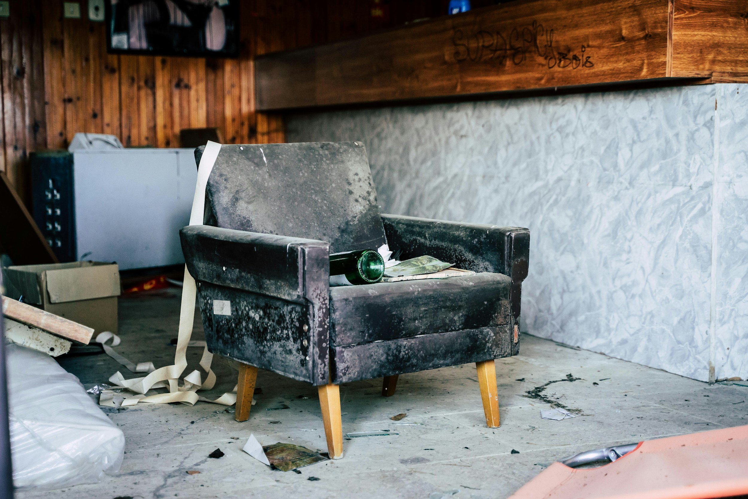 A worn, dusty armchair with wooden legs in a cluttered and messy room, surrounded by trash, debris, and miscellaneous objects, with wooden and stone walls and a damaged floor.