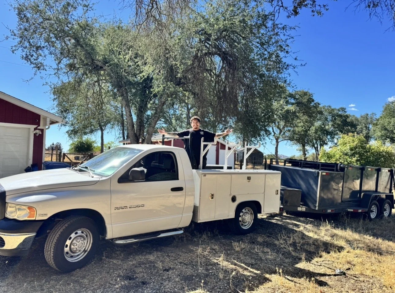 Person standing through the side window of a white service truck with an extended bed, attached to a black trailer, in a rural area with trees and a house nearby.