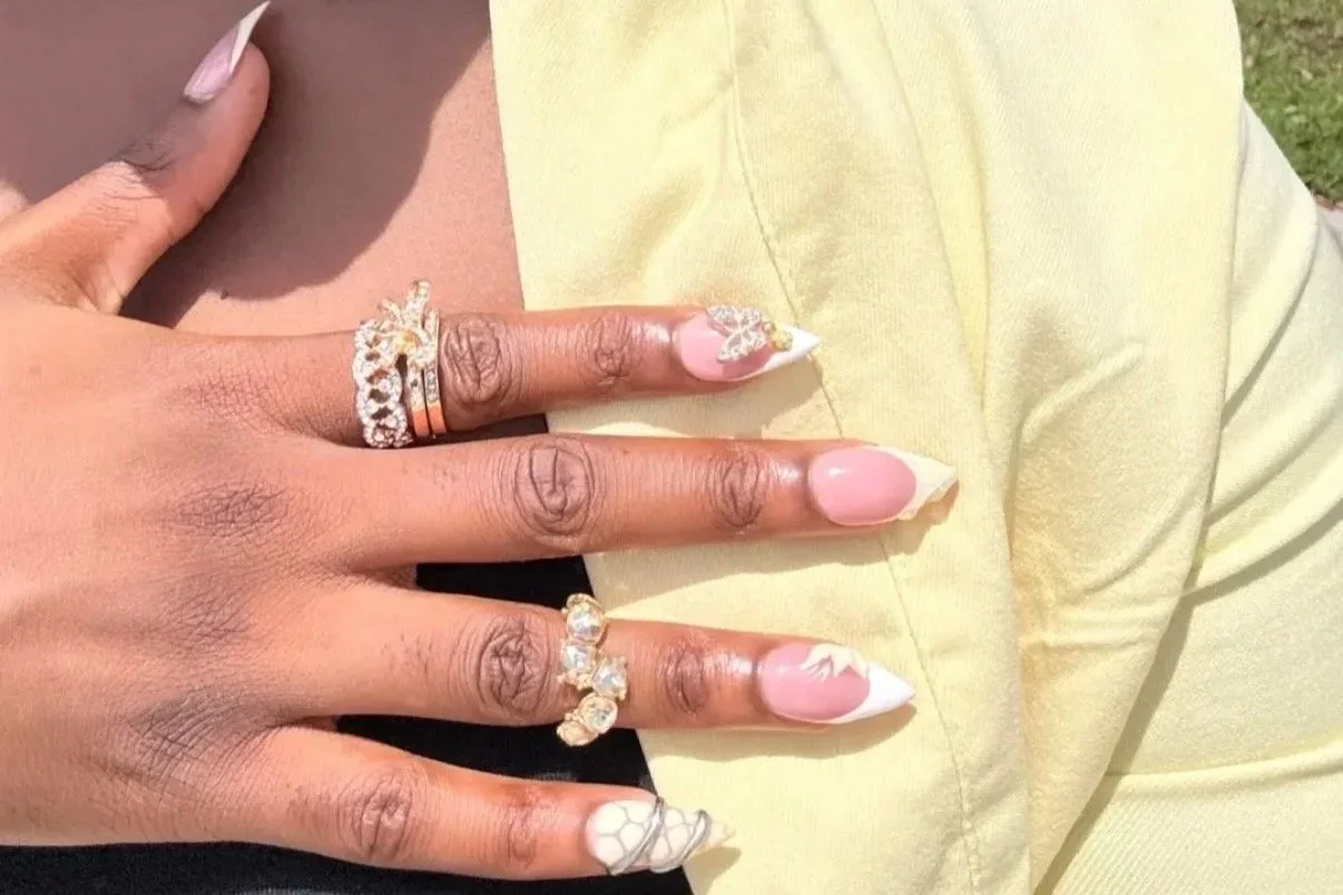 Close-up of a hand with decorated nails, multiple rings, and a person wearing a yellow outfit with a black top underneath.